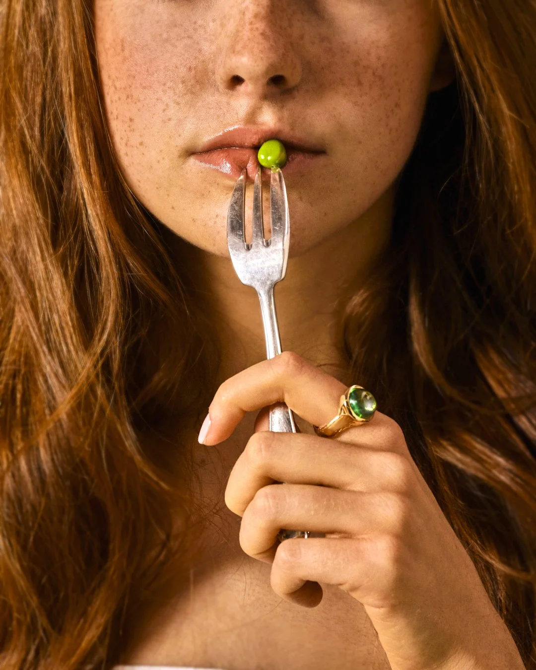 Une femme aux cheveux roux tient une fourchette avec un pois vert entre ses dents, portant une bague verte. 