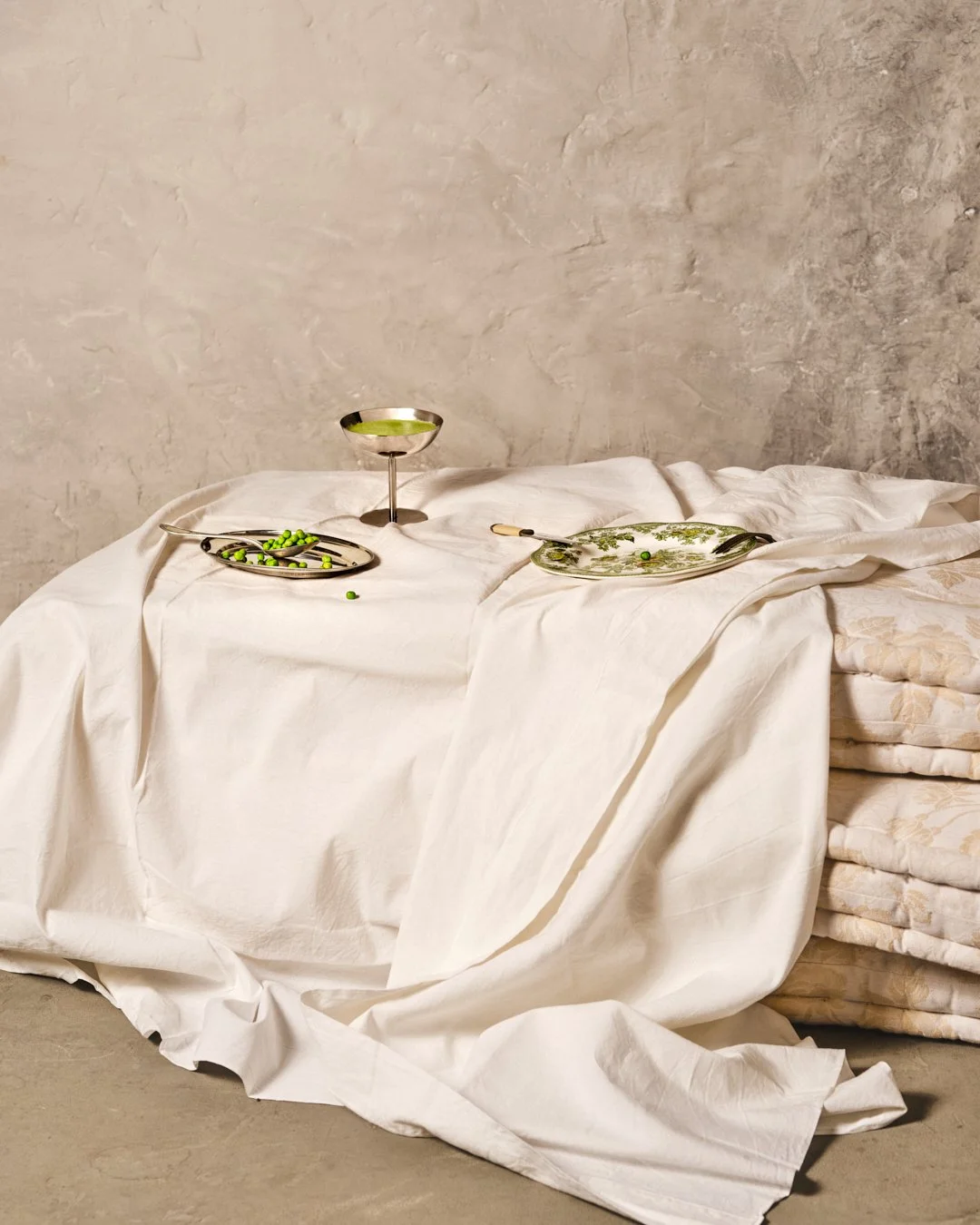 Une table recouverte de draps blancs avec des assiettes, un verre et des pois verts, côté lit avec des matelas empilés, devant un mur en béton.