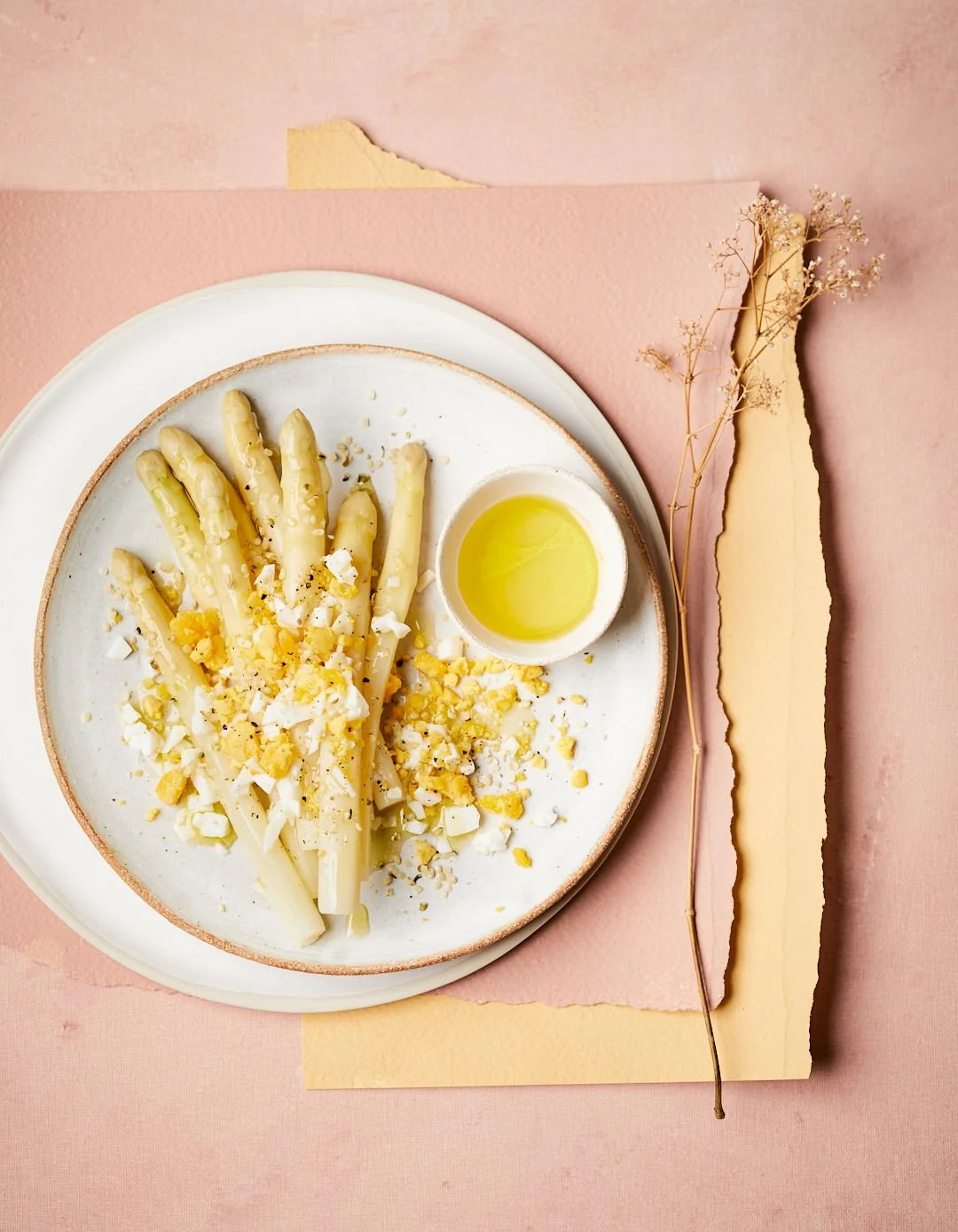 Asperges blanches avec œufs durs, copeaux de fromage, et huile d'olive dans un bol, présentées sur une assiette blanche sur un fond de papier rose et beige.