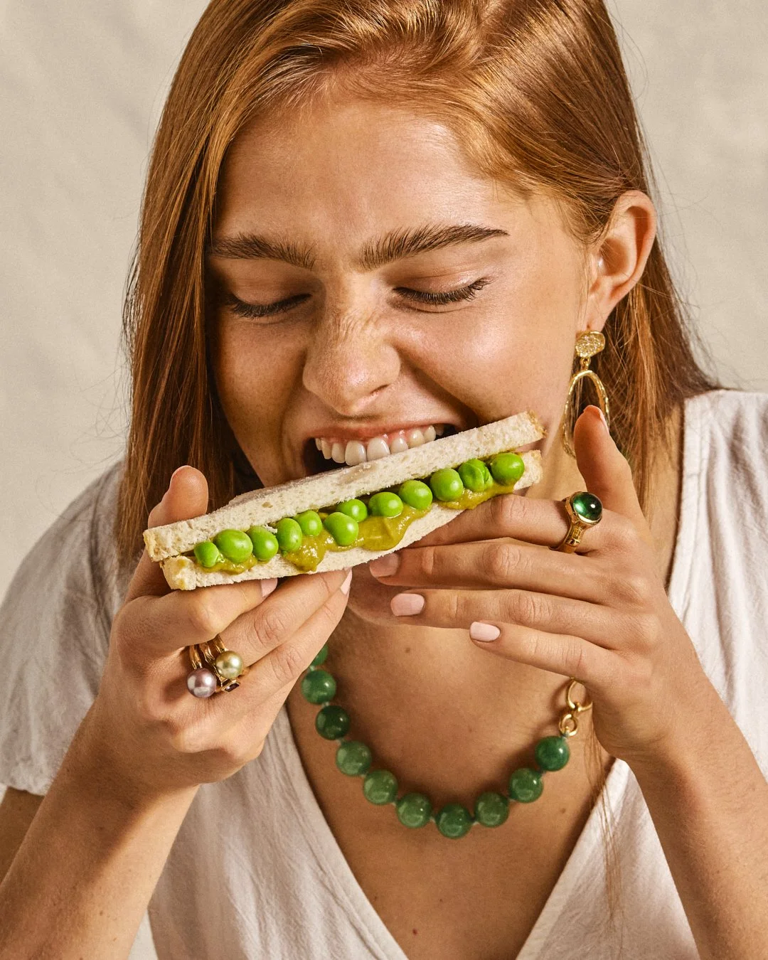 Une femme aux cheveux roux, portant des bijoux en or et des perles vertes, tient un sandwich avec des pois verts et du moutarde.