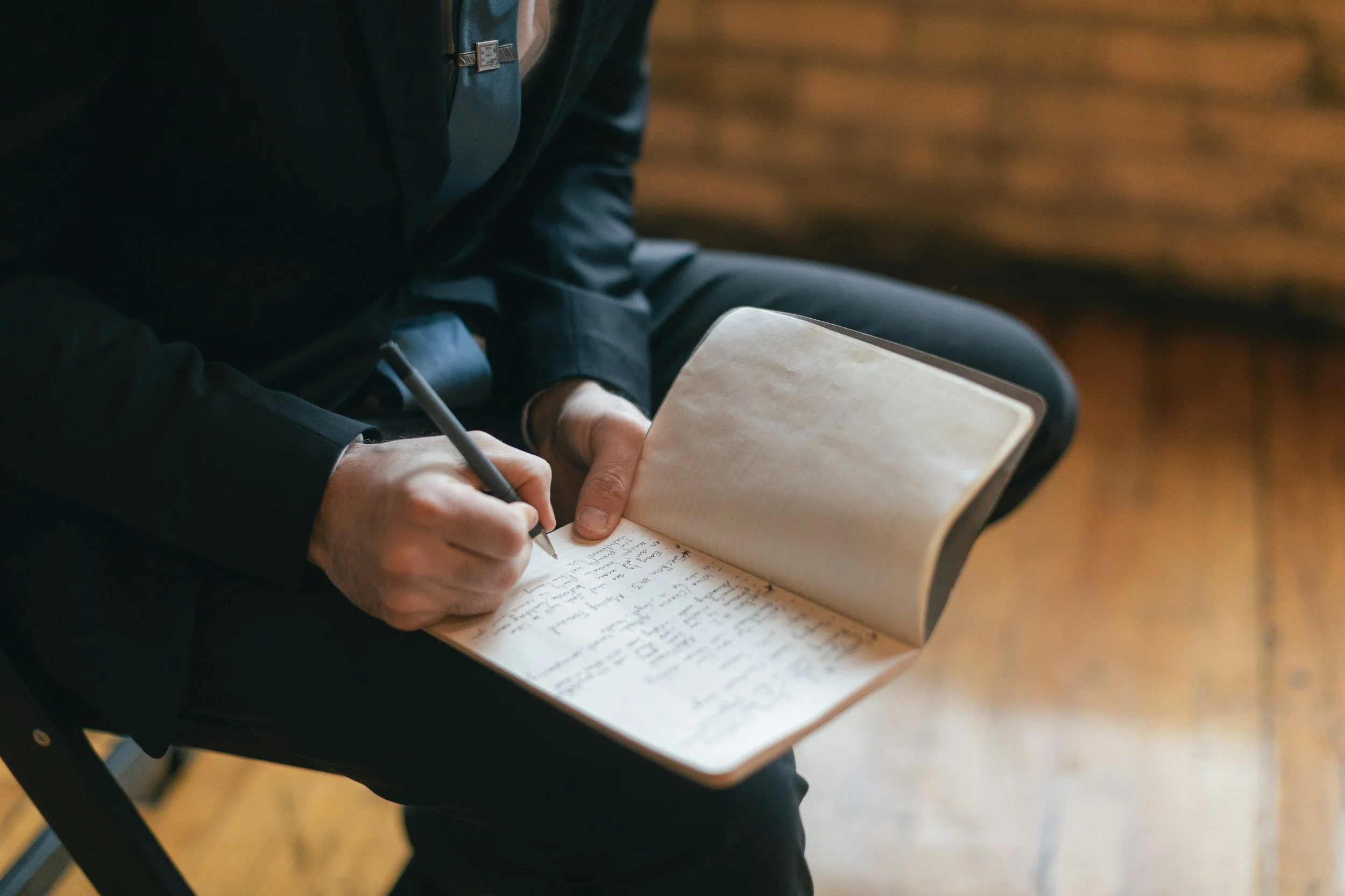 Person seated, writing notes in a notebook with a pen, the notebook is on their lap, and they are wearing a dark jacket and pants.