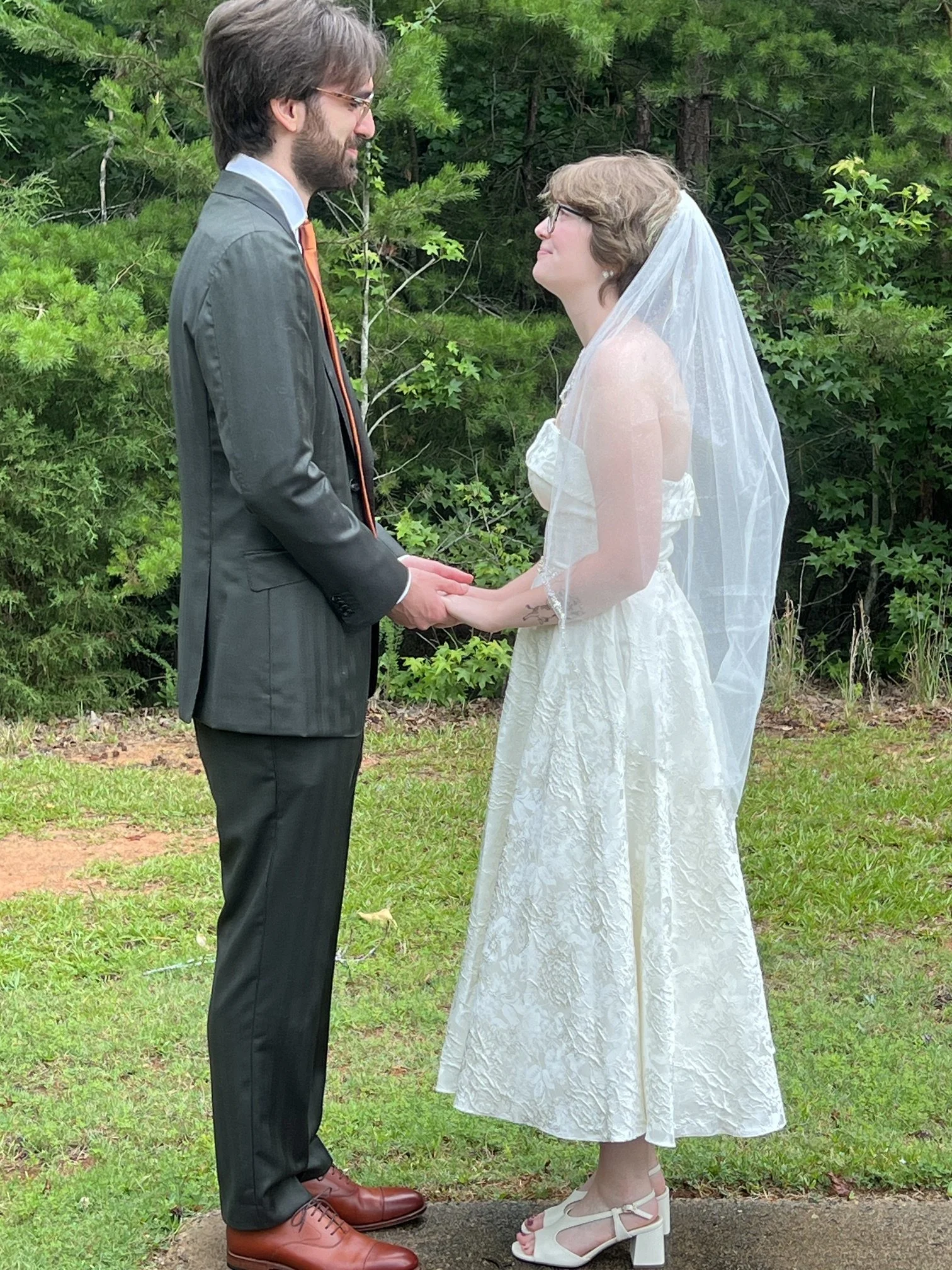 A bride and groom holding hands face to face outdoors in a wooded area, dressed in wedding attire. The groom wears a gray suit with an orange tie, and the bride wears a white lace wedding dress with a veil and white heels.