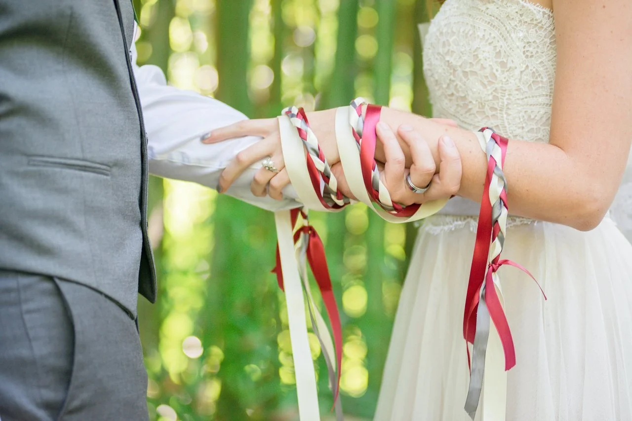 Close-up of a wedding ceremony with the bride and groom holding hands, adorned with red and cream ribbons in a lush green outdoor setting.