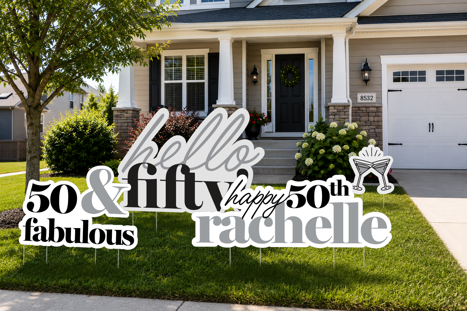 Decorative birthday sign on a lawn celebrating a 50th birthday for someone named Rachel, with phrases like 'hello,' 'fifty,' 'happy,' 'fabulous,' and '50th' surrounding a pair of clinking glasses graphic, in front of a house with gray siding, white trim, and a black front door.