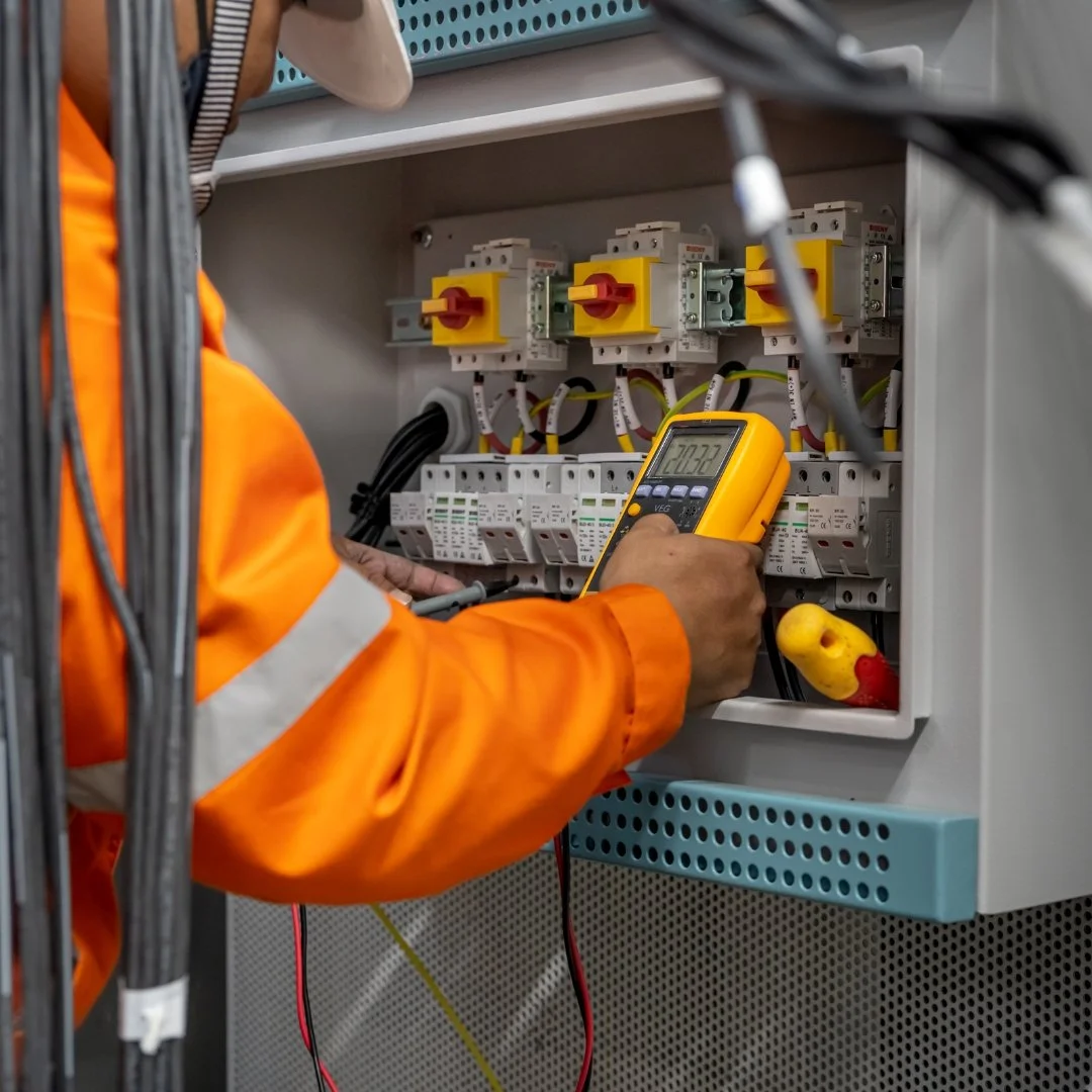 An electrician in an orange safety jacket using a multimeter to test an electrical panel with circuit breakers.