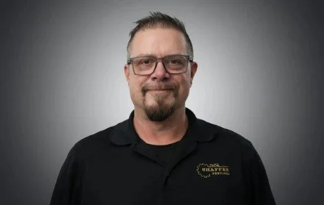 A man with glasses and a beard, wearing a black collared shirt, standing against a gray background.