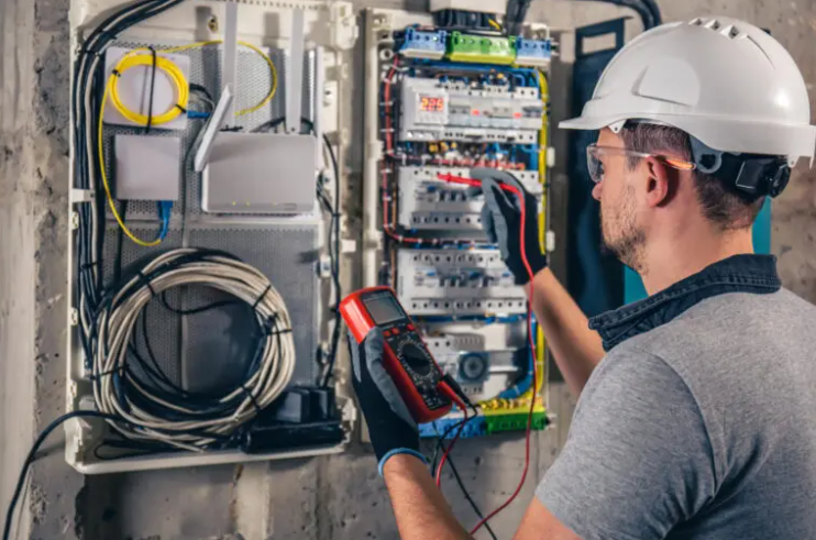 Electrician wearing a white helmet using a multimeter to test wiring inside an electrical panel.