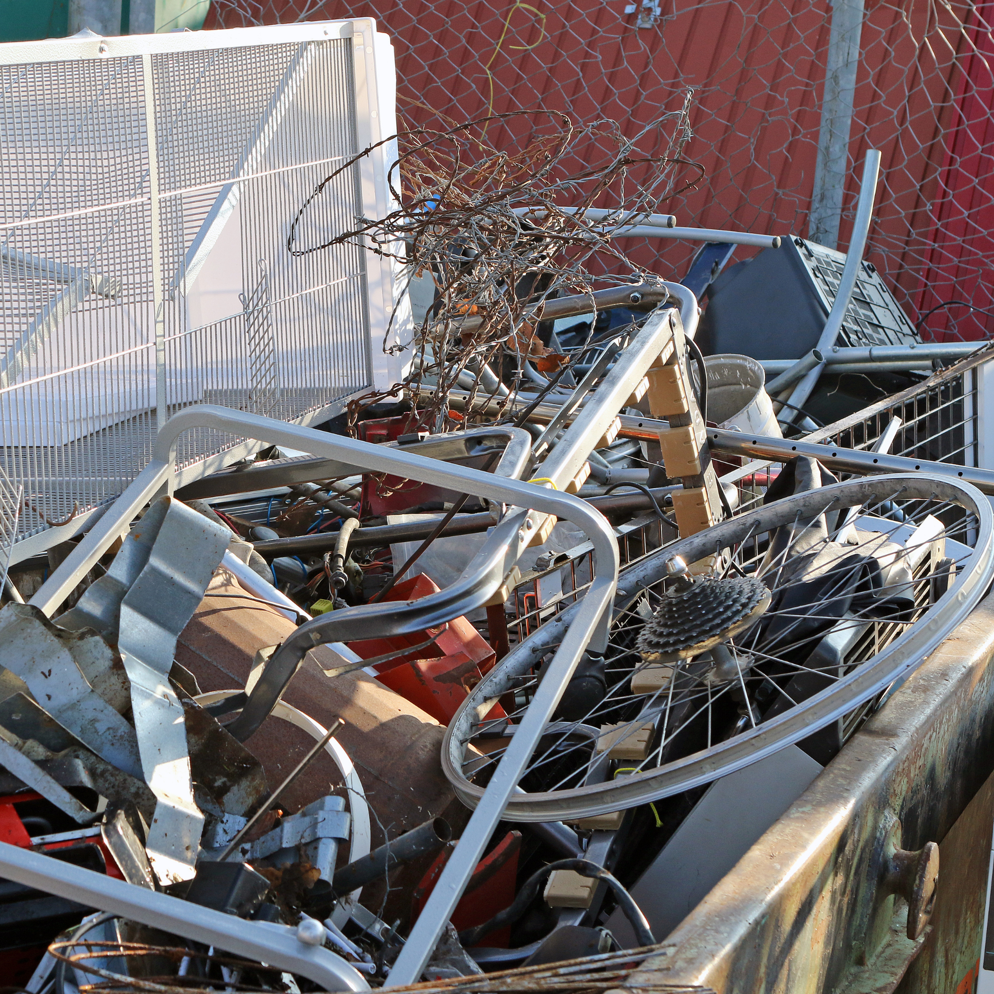 A pile of discarded metal frames, tangled wires, and scrap debris outdoors next to a chain-link fence and red wall.