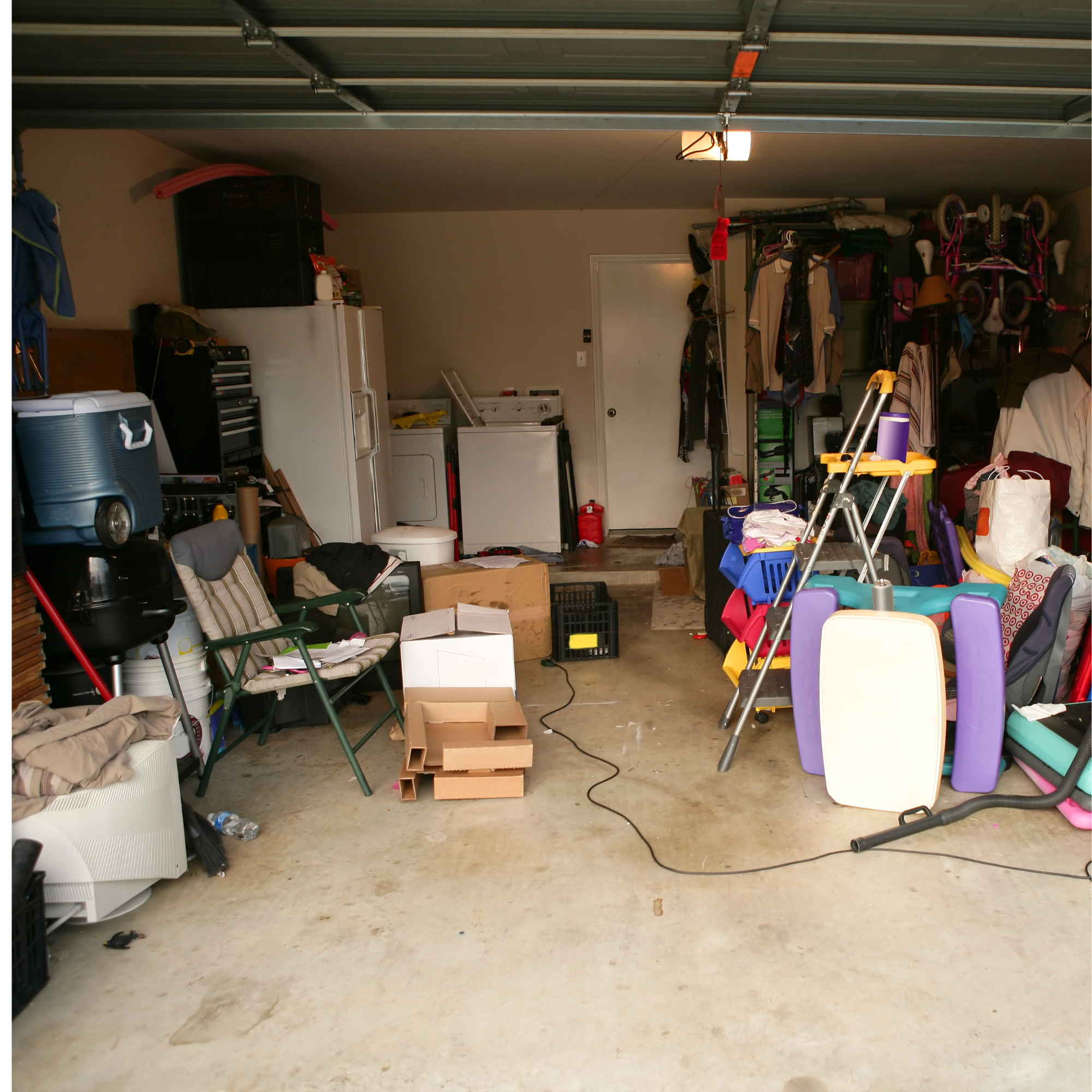 A cluttered garage with various household items, including chairs, a cooler, boxes, a washing machine, a bicycle, and miscellaneous household objects. The garage has a concrete floor with some stains and an open ceiling with exposed beams.
