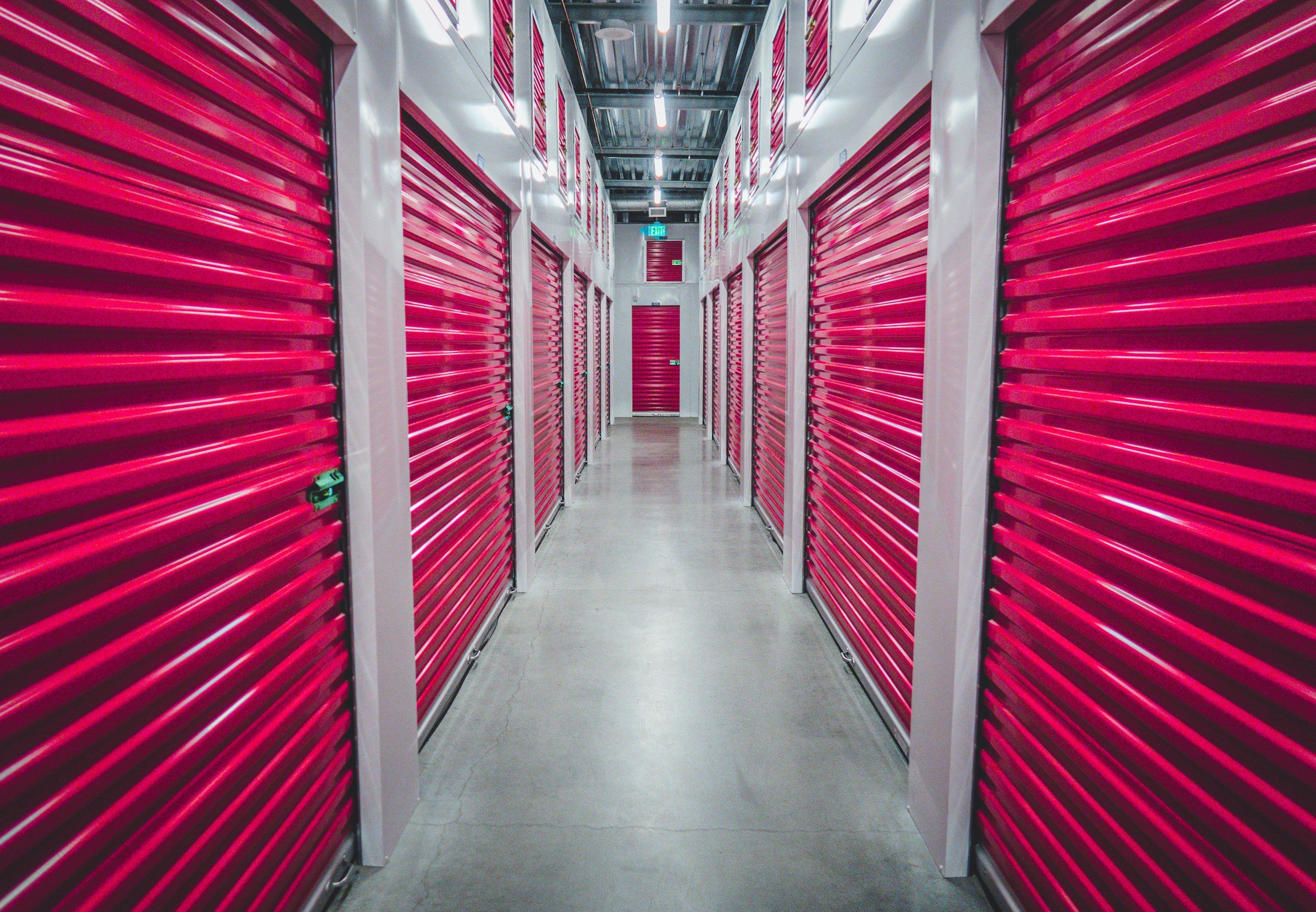 A hallway in a storage facility with multiple red storage unit roll-up doors on either side.