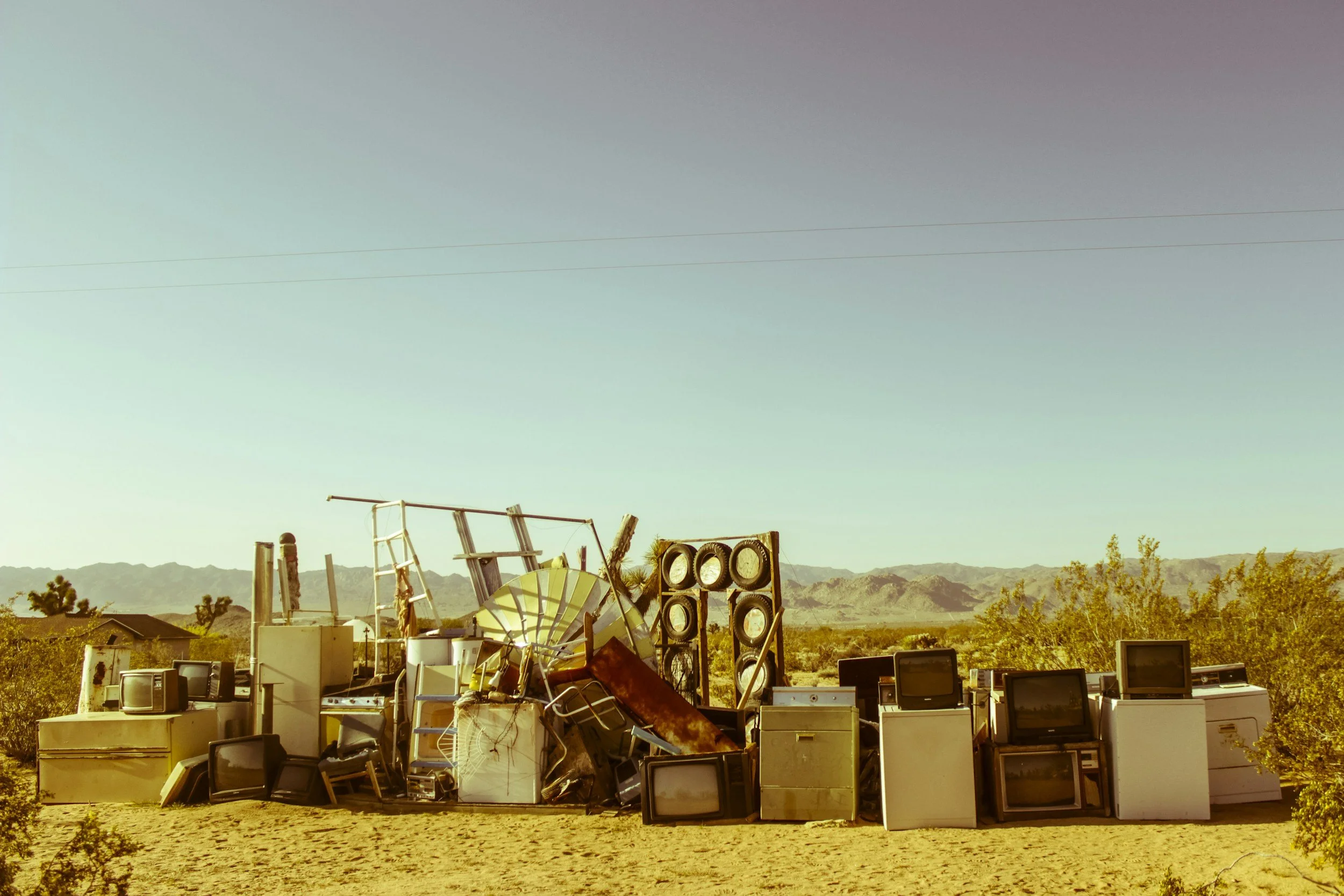 A large collection of old electronics, including many CRT monitors and TVs, stacked haphazardly outdoors with a desert landscape and mountains in the background.