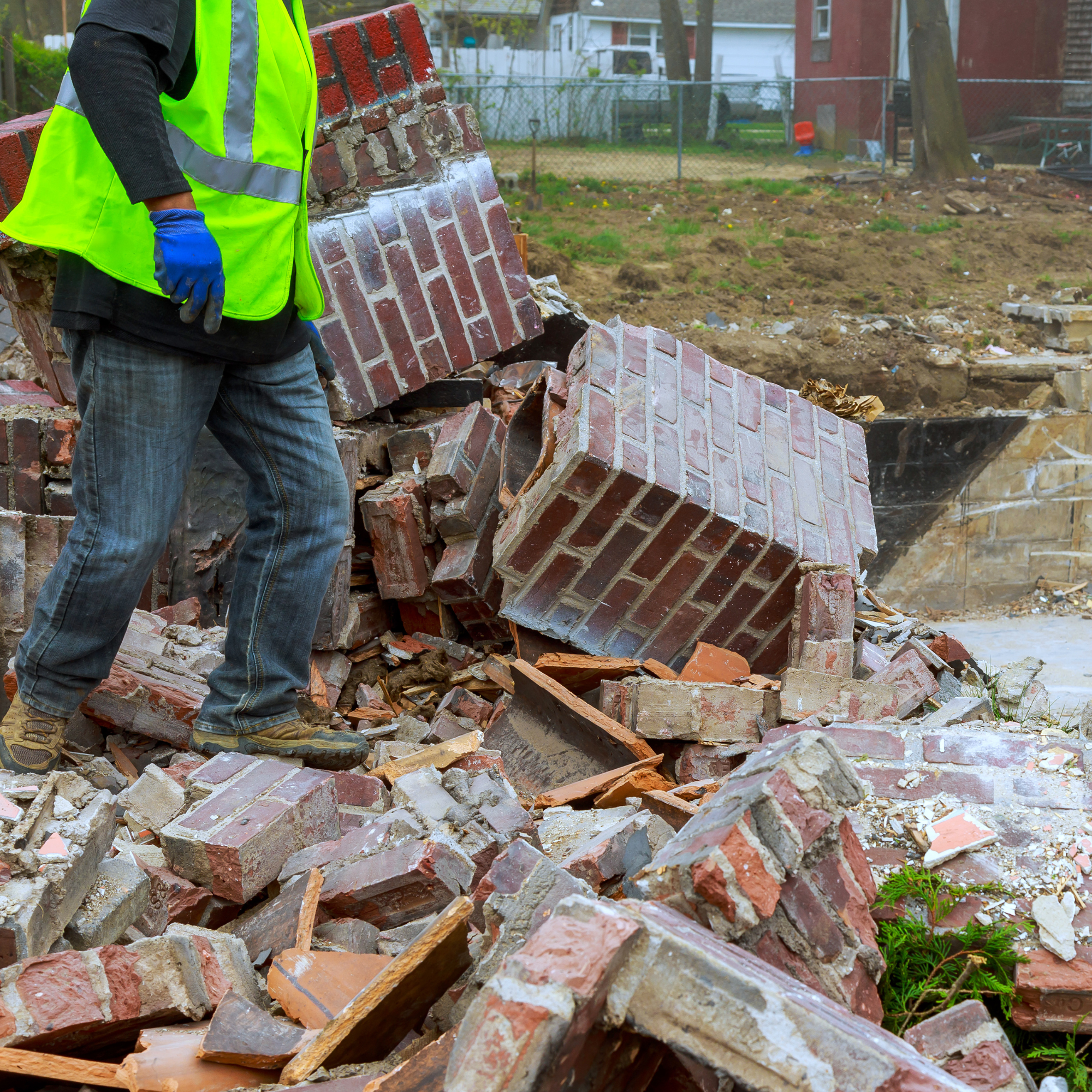 A person wearing a high-visibility vest, blue gloves, and sturdy boots is demolishing a brick wall in a residential backyard, with broken bricks and debris on the ground.