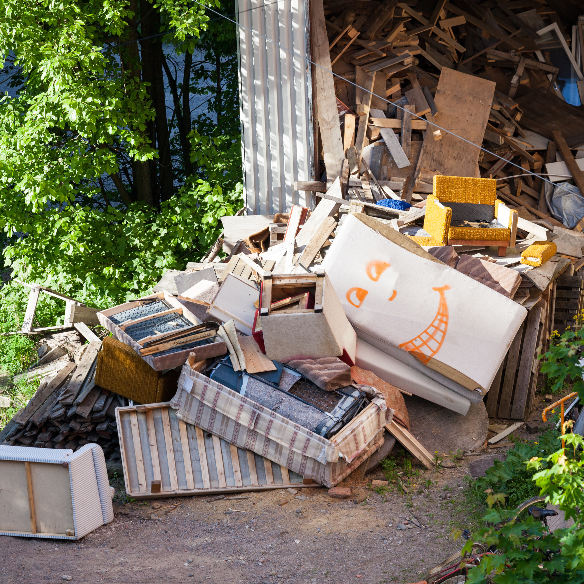 A cluttered outdoor area with various pieces of wooden furniture, furniture parts, and construction debris. There are green trees on the left side and a dirt ground in the foreground.