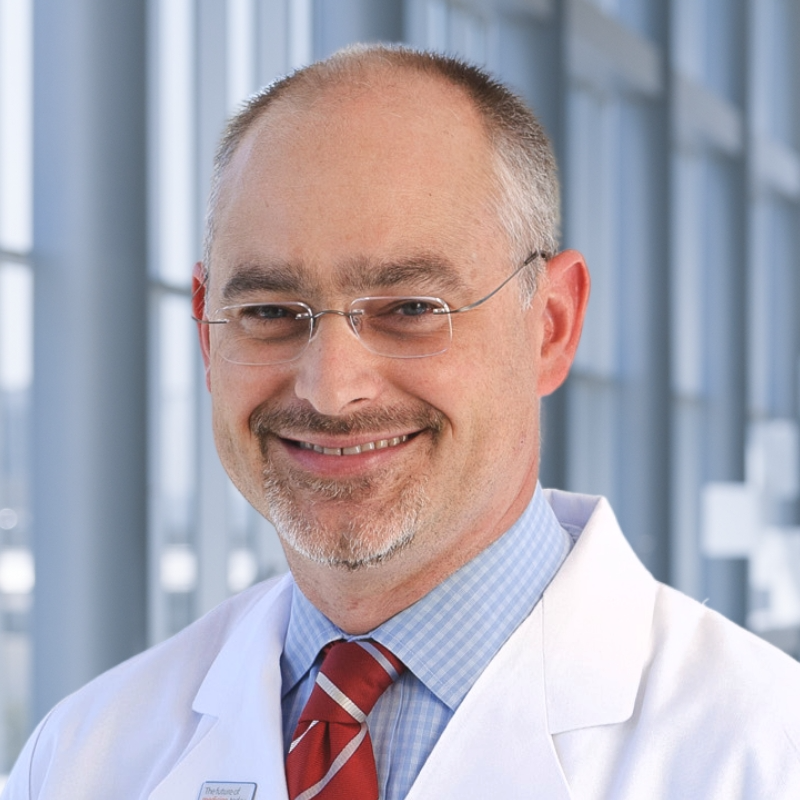 A smiling male doctor with glasses, a beard, and a mustache, wearing a white coat, light blue shirt, and red striped tie, standing in a modern medical building with large windows in the background.