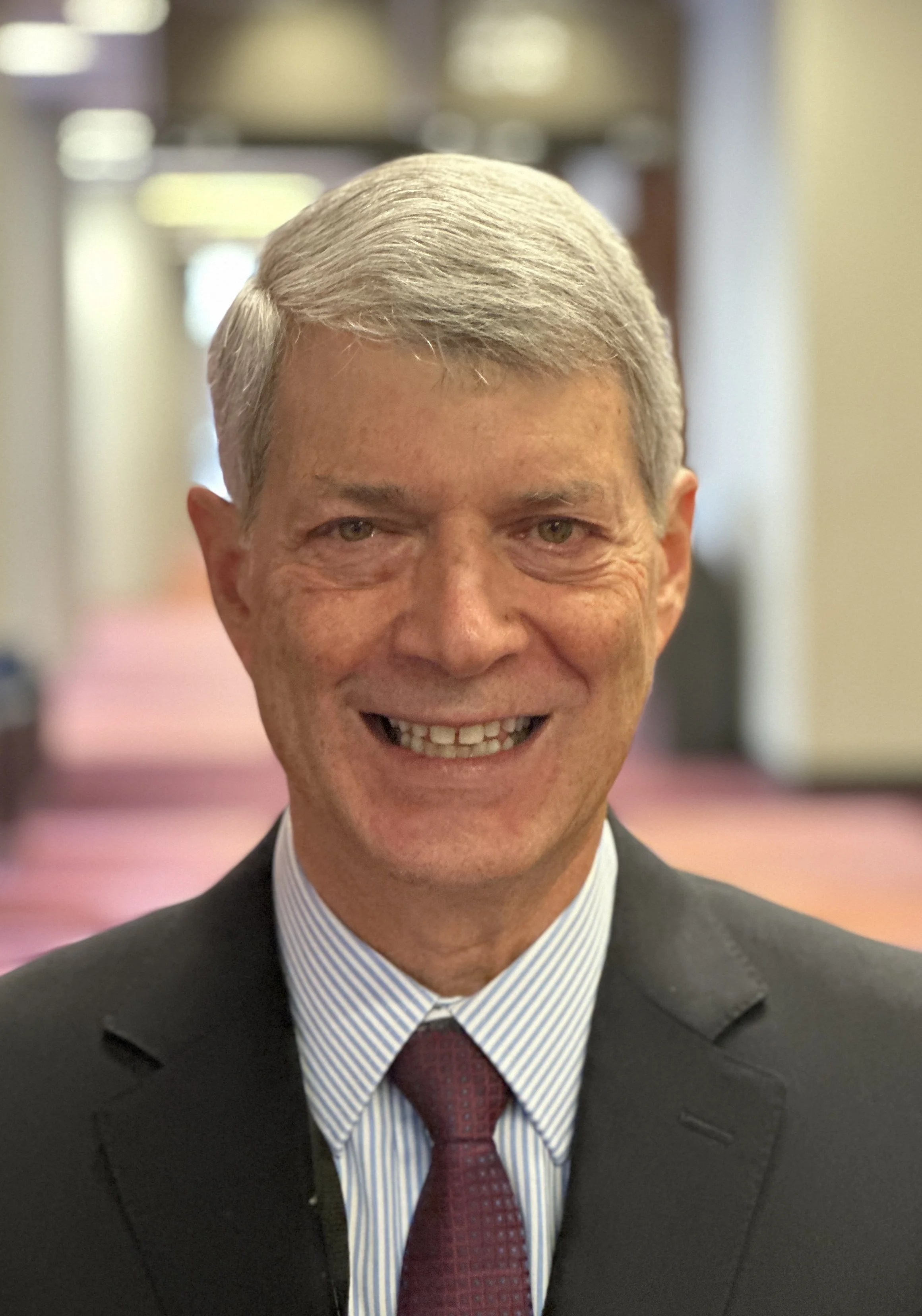 Close-up of a smiling middle-aged man with gray hair, wearing a dark suit, a light blue striped shirt, and a red tie, at a formal event in an indoor setting.
