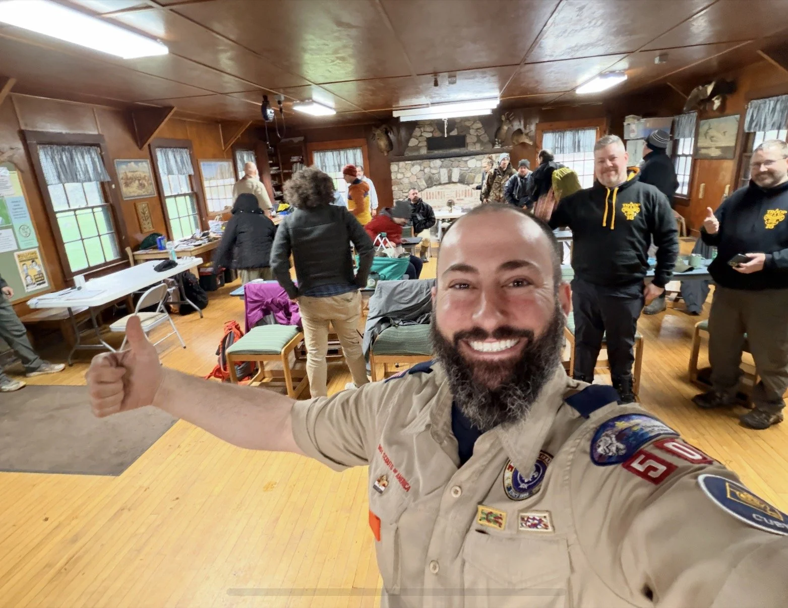 A smiling man with a beard in a beige Boy Scout uniform taking a selfie in a room filled with people, some standing and sitting, with wood-paneled walls, windows, and a stone fireplace in the background.