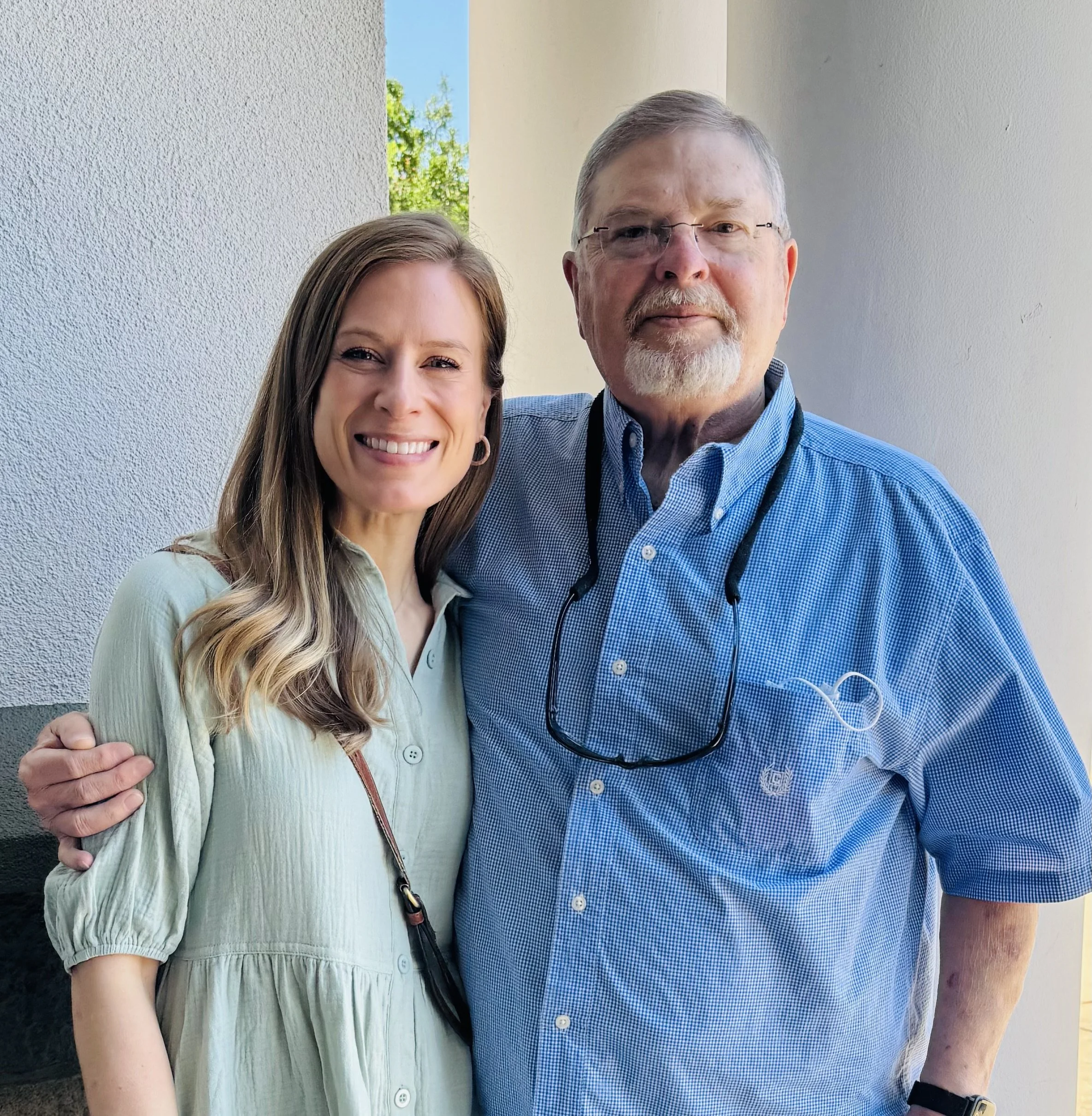 A man and woman standing outside near a white wall, smiling. The woman has long, wavy hair, wearing a light green dress, and the man is older with gray hair, glasses, wearing a blue checkered shirt and a black lanyard.