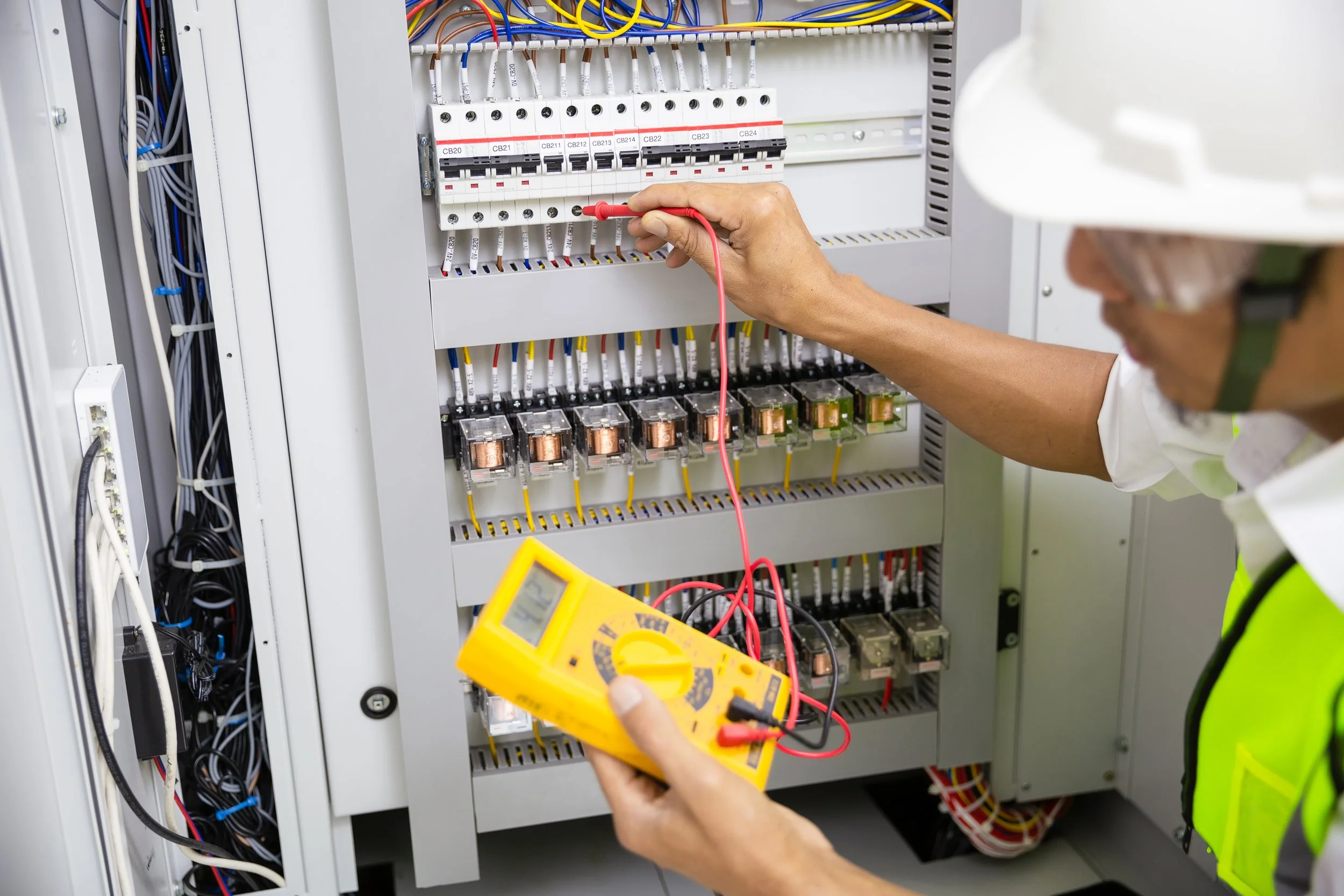 Technician testing electrical panel with a multimeter in an industrial or electrical setting.