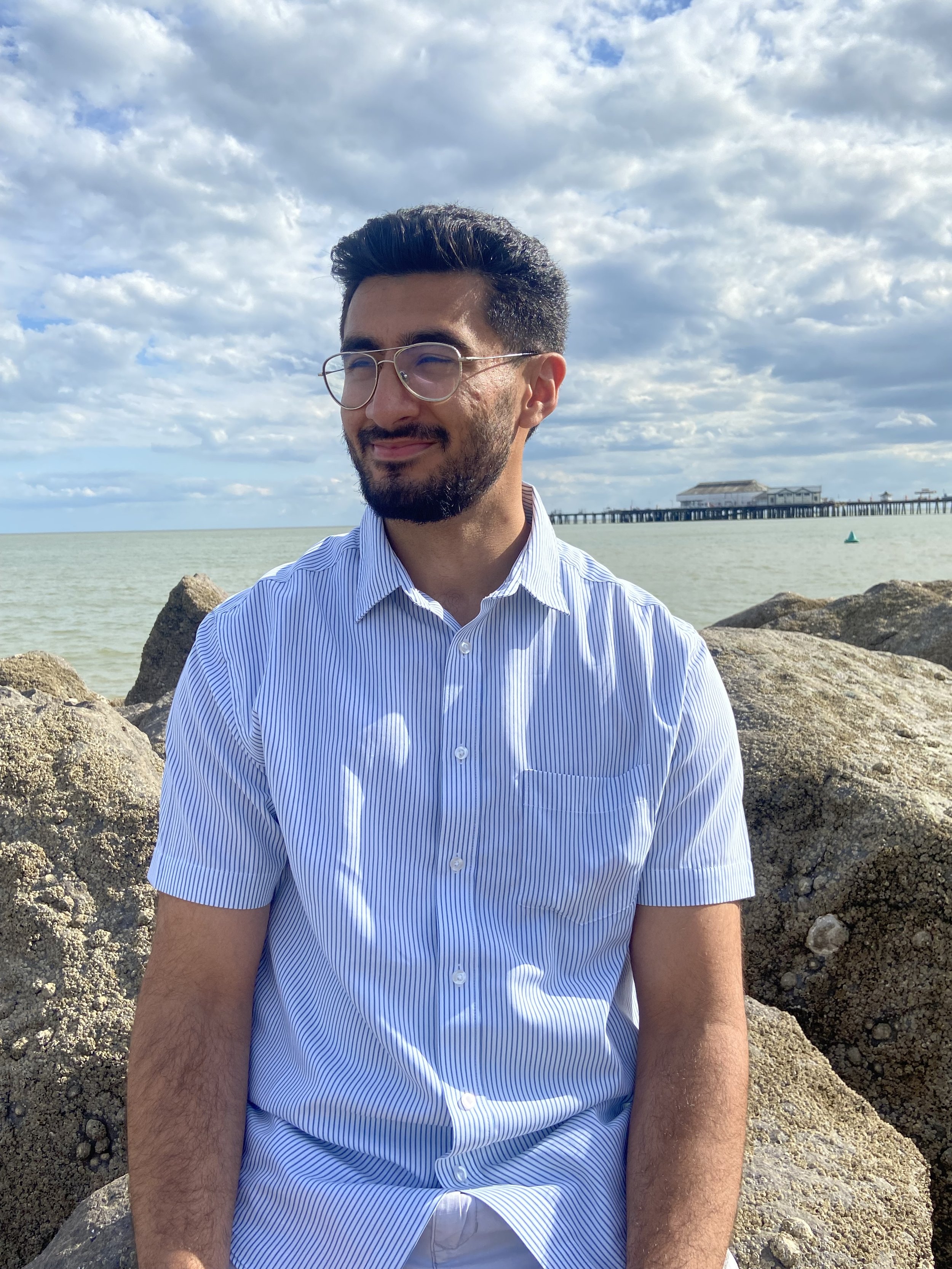 A man with glasses and a beard, wearing a blue striped shirt, sitting on rocks near the water at a pier, with clouds and a cloudy sky overhead.