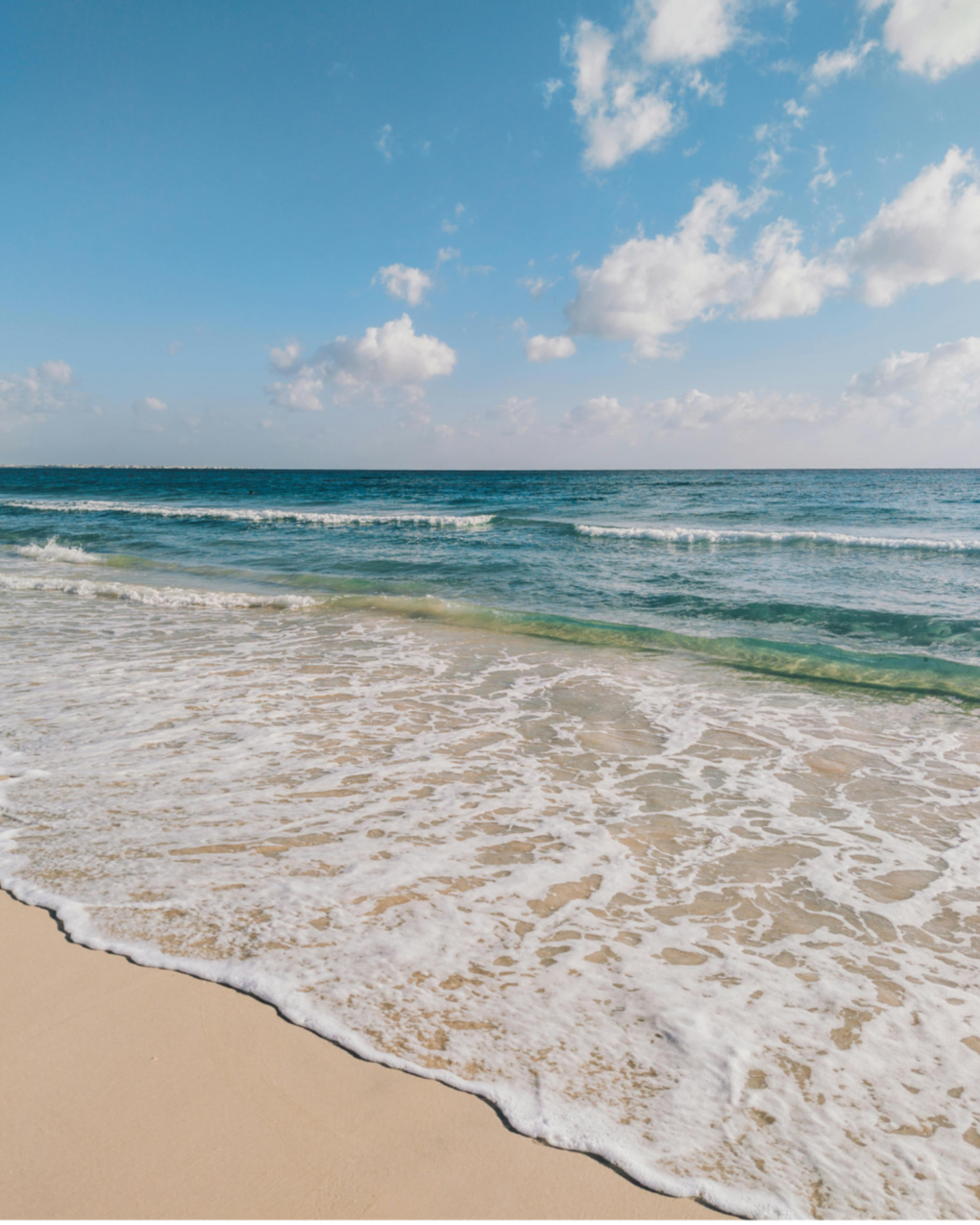 A sandy beach with gentle waves and a clear blue sky with scattered clouds.