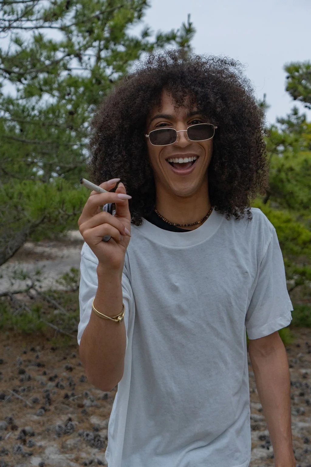 A person wearing sunglasses and a white tee holding a pre-roll in the woods
