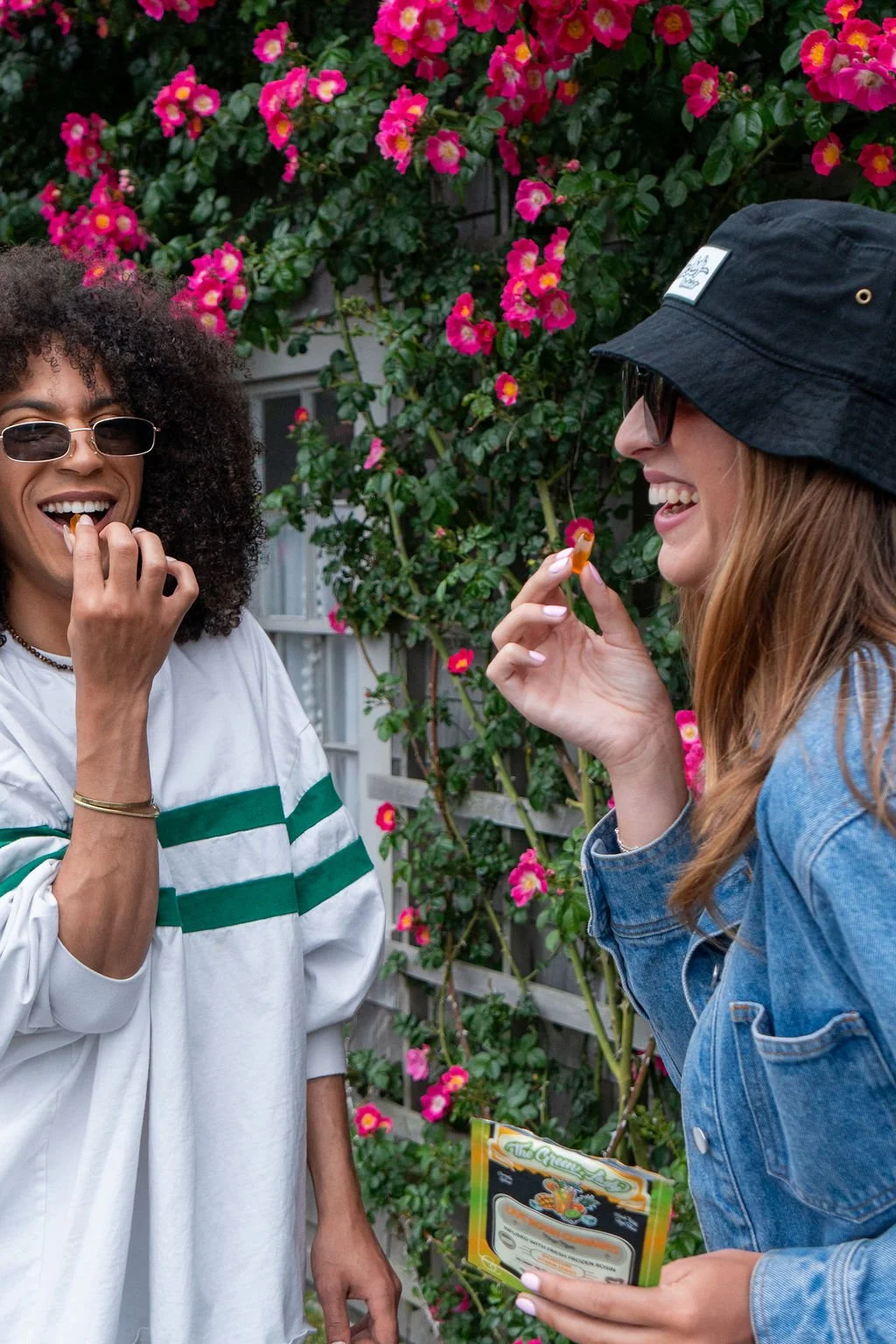 Two friends eating gummies and smiling in front of a wall of flowers