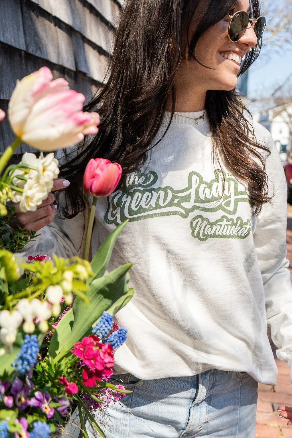 A young woman standing with flowers wearing a white crewneck sweatshirt with The Green Lady Nantucket logo