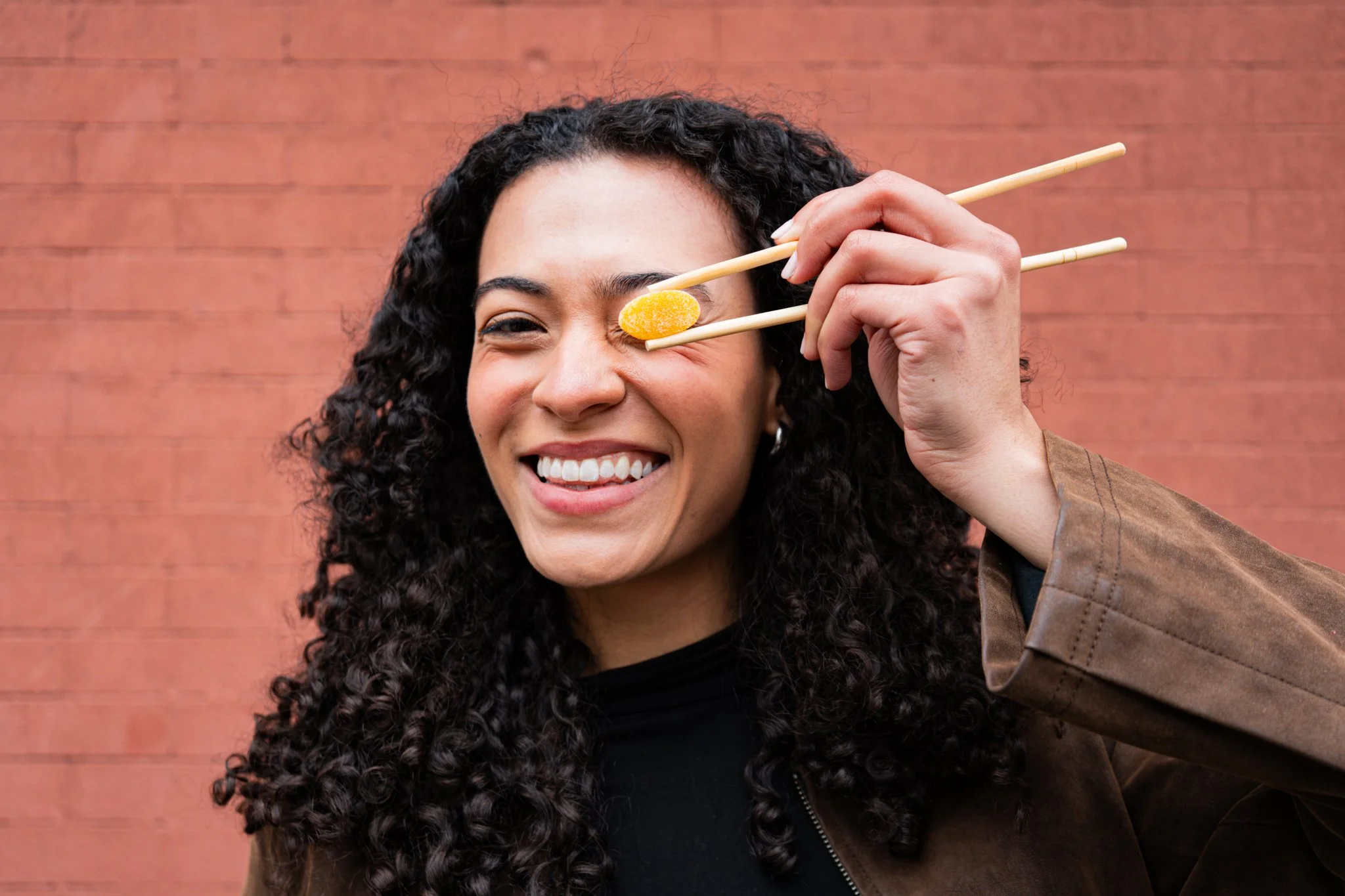 A young woman standing in front of a brick wall holding a gummy with chopsticks in front of her eye