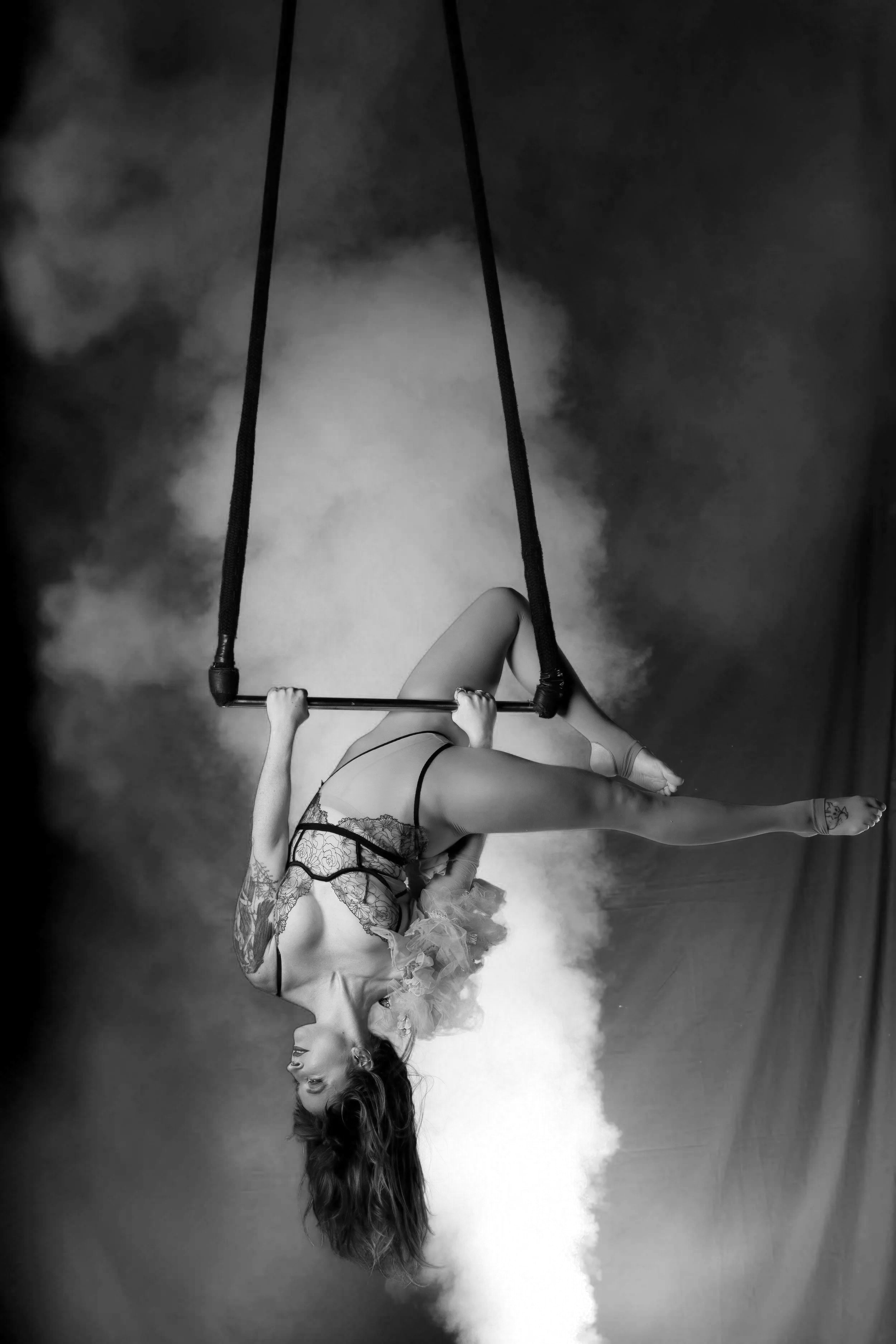 A woman performs an aerial silk routine, hanging upside down from black silk fabric against a background with pink and blue smoke or fog.