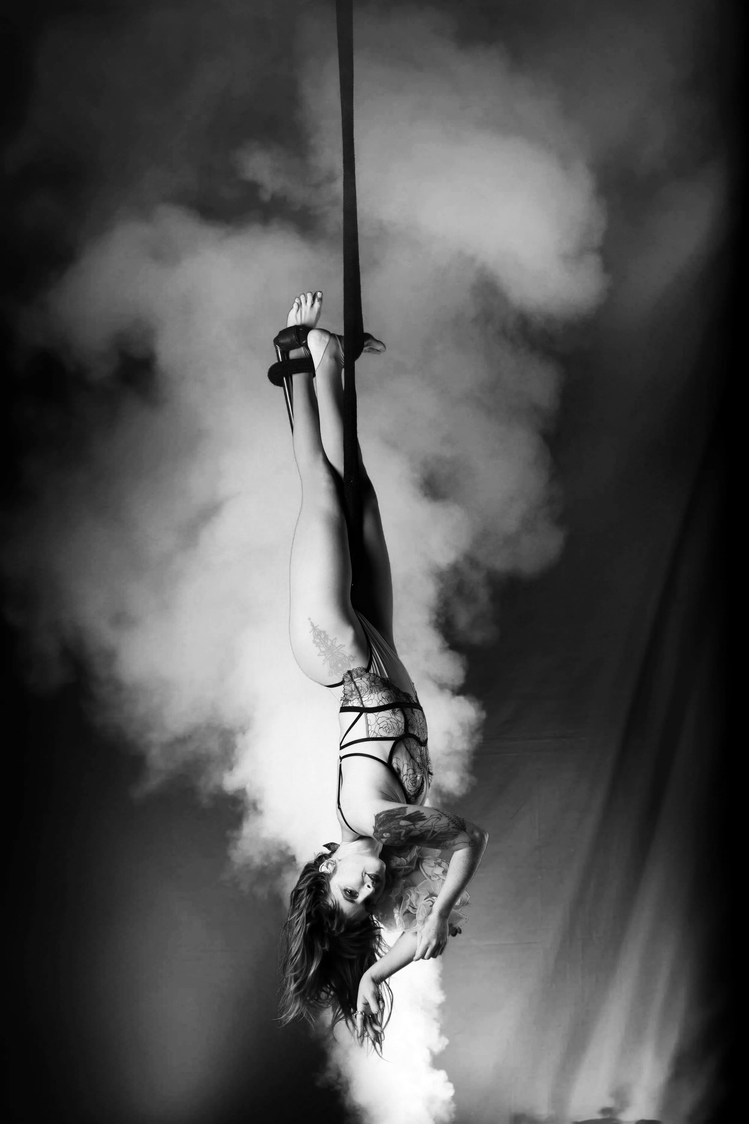 A woman performing an aerial pose, hanging upside down from a black strap, with a colorful smoke background in purple and pink hues.