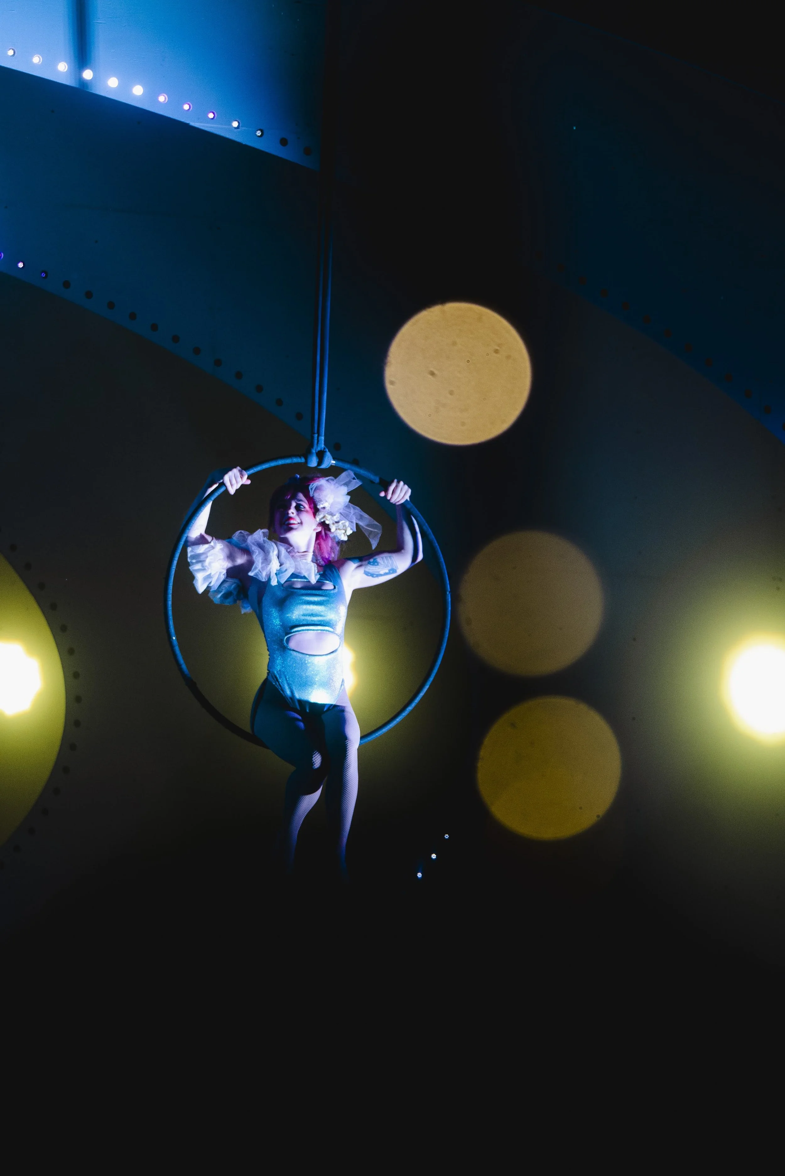 A female performer in a shiny costume with puffy sleeves and fishnet tights is hanging on a black aerial hoop, smiling. The background features circular light patterns and yellow spotlights.