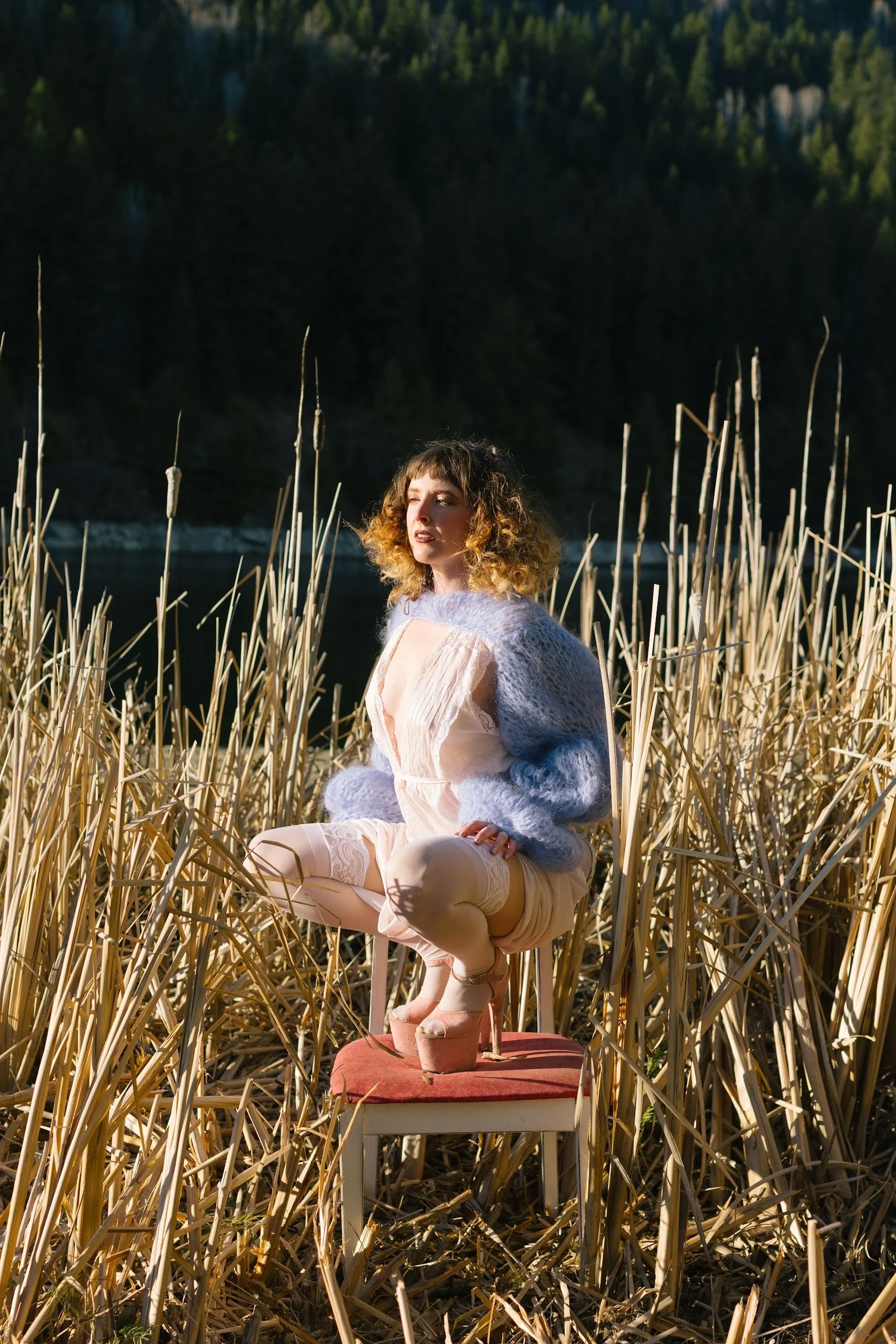 A woman with curly hair wearing a pink dress, lace thigh-high stockings, pink high heels, and a light gray fur shawl, sitting cross-legged on a padded chair in a marshy area with tall reeds in front of a body of water and green forested hills in the 