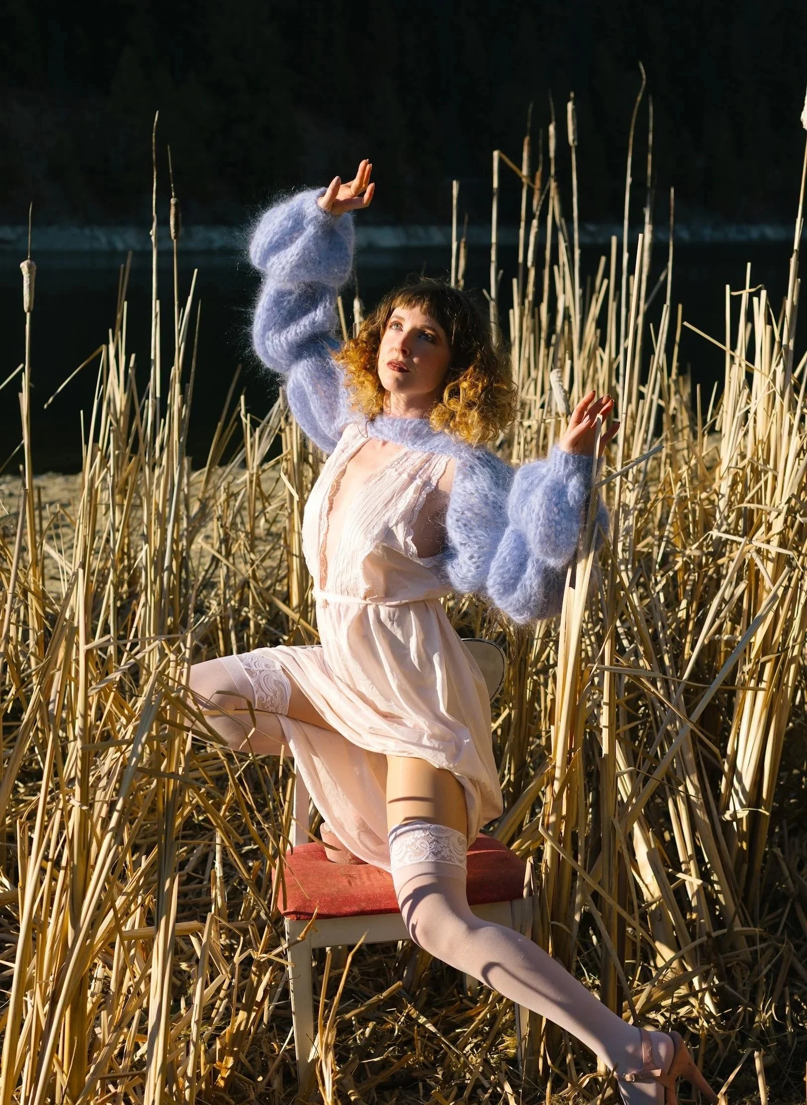 A woman with curly hair in pale pink dress, thigh-high stockings, and lavender fuzzy sweater, kneeling among dry tall reeds near water at sunset, raising her arm with fingers extended.