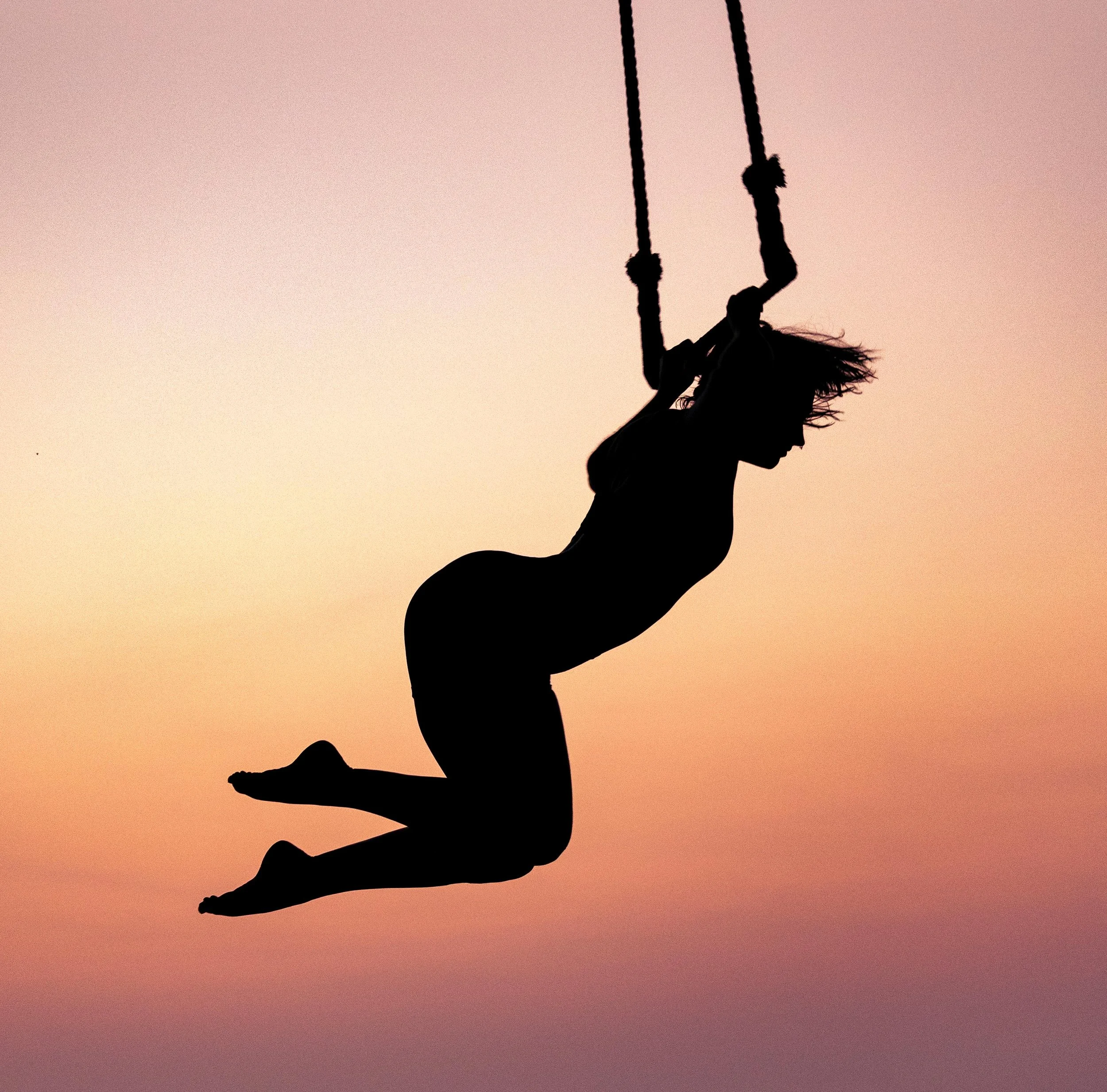 Silhouette of a girl with flowing hair swinging on a swing, against a gradient sky at sunset or sunrise.