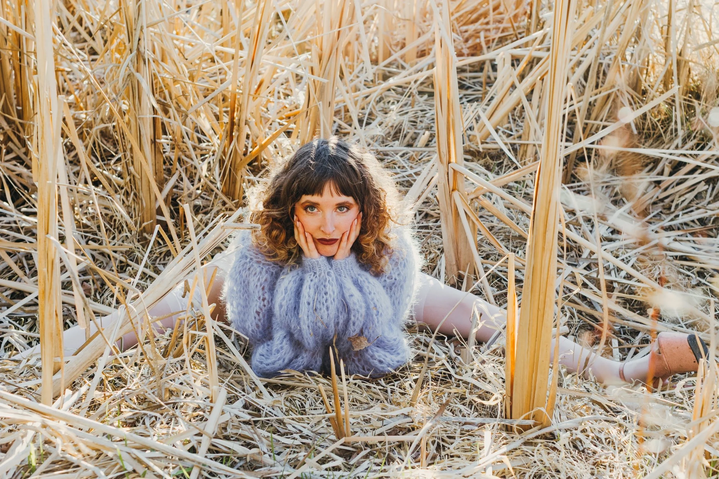 A woman with curly hair and wearing a fluffy gray sweater is lying on the ground in a field of dry wheat stalks, with her head resting in her hands and looking directly at the camera.