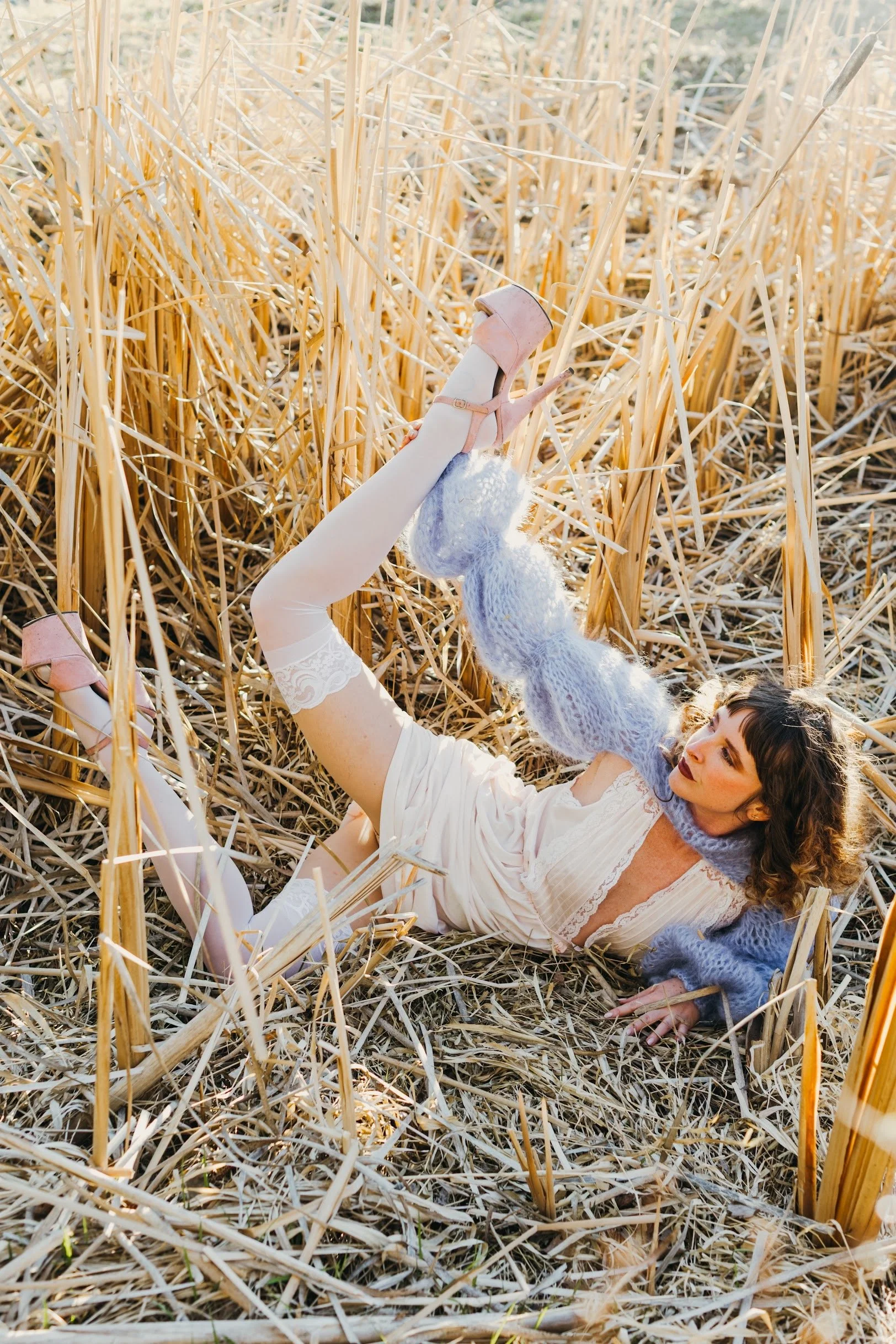 A woman lying on her back in a wheat field, wearing a white lace dress, lavender sweater, and pink high-heeled shoes, with her legs raised and crossed in the air.