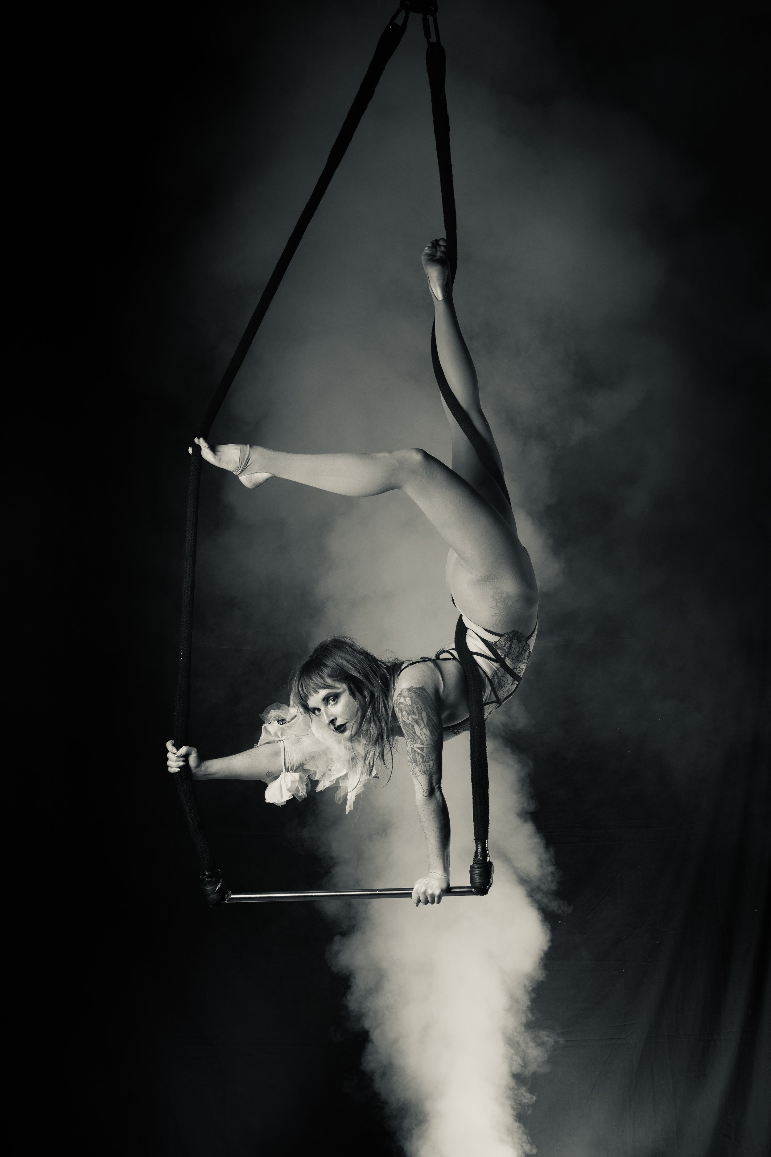 A female aerial performer in a black and white photograph poses on a trapeze, with her body upside down and legs extended upwards. She has tattoos and wears a light-colored tutu, and her face has dark makeup and expressive eyes. Smoke or mist billows below her, creating a dramatic scene.