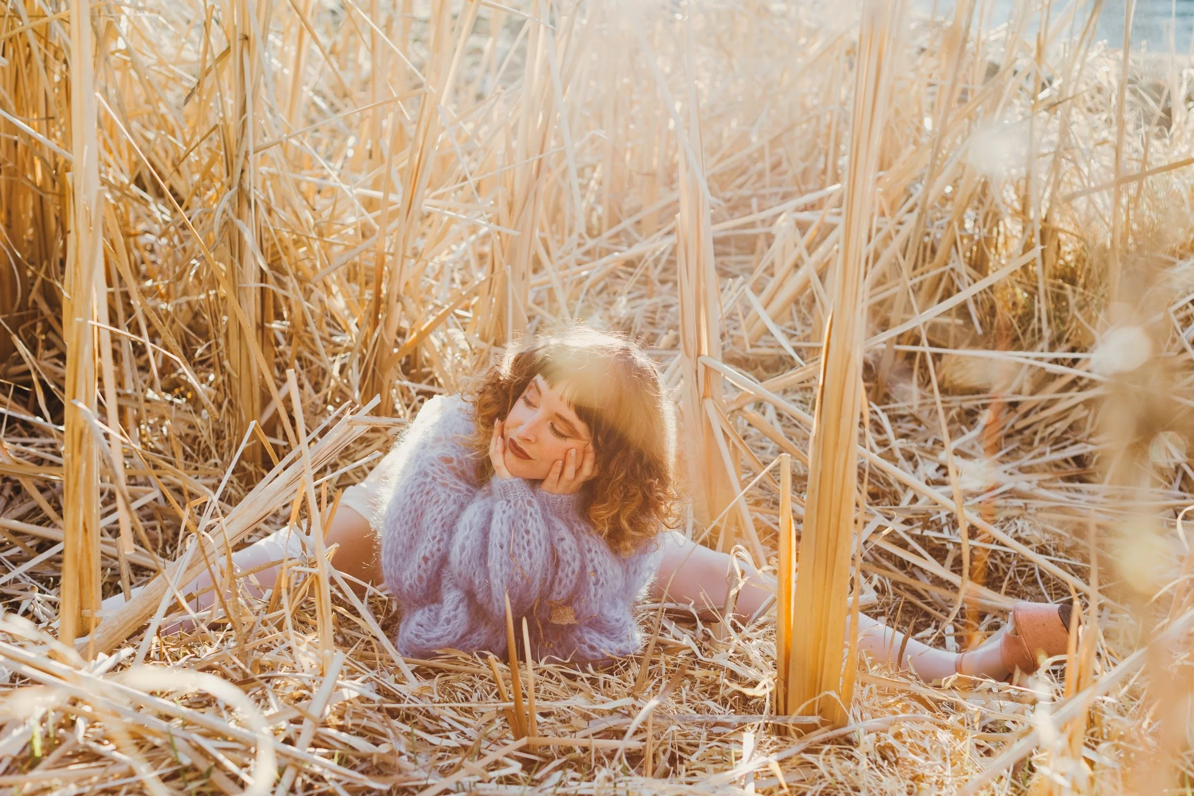 A woman with curly hair and a sweater sitting among dried tall grass, with sunlight shining through, appearing relaxed.