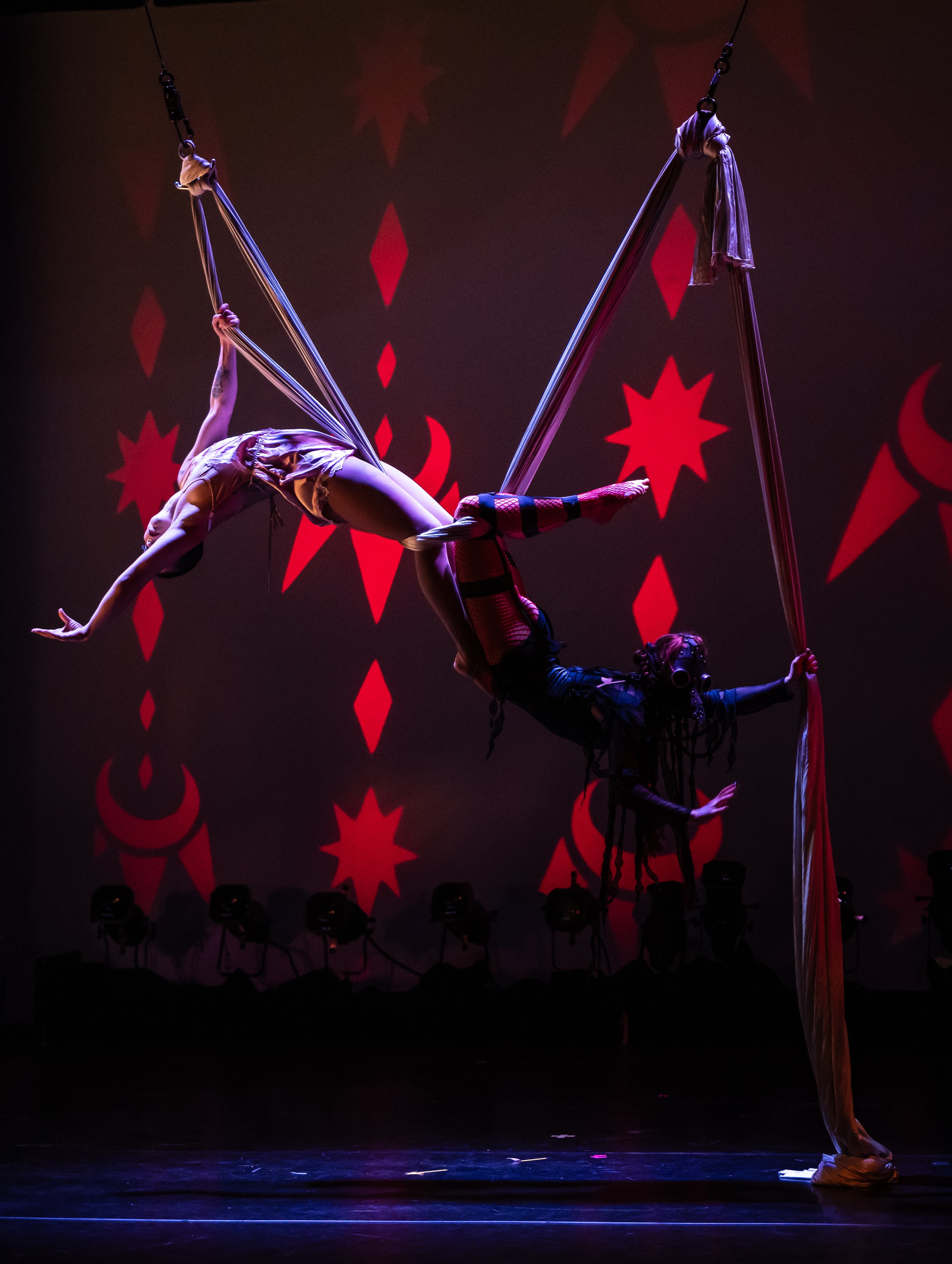 Two aerial acrobats perform on a stage with red star and crescent moon decorations in the background. One acrobat is hanging upside down by their legs from silk ribbons, while the other supports with their hands and feet. The stage is dark with spotl