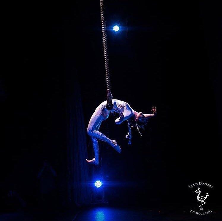 A performer in a shimmering leotard hanging upside down from a rope during a dance or circus performance, illuminated by stage lights with a dark background.