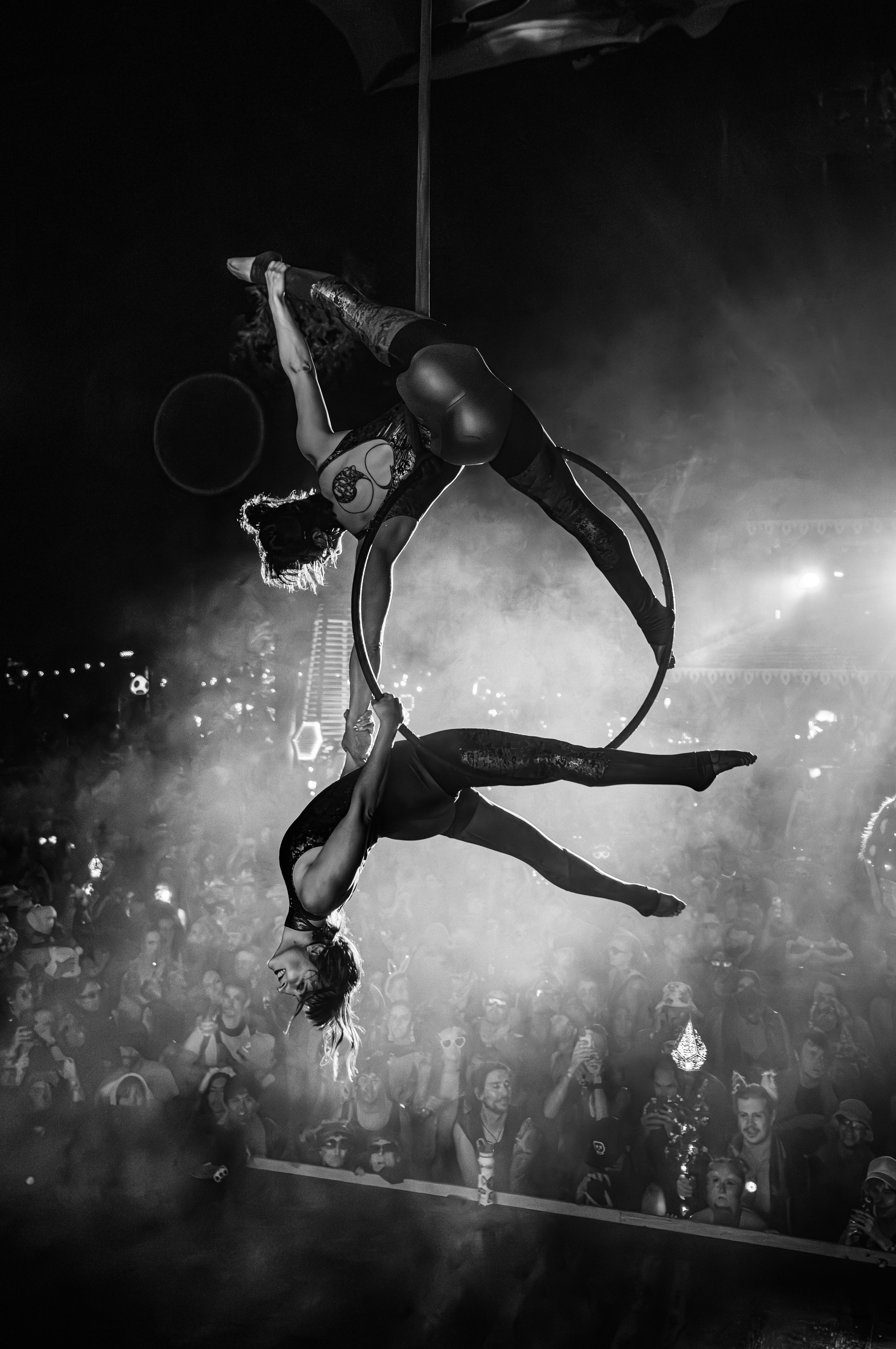 Two acrobats perform a balancing act with hoops in a circus arena, surrounded by an audience in the background. The image is in black and white, emphasizing the dramatic lighting and shadows.