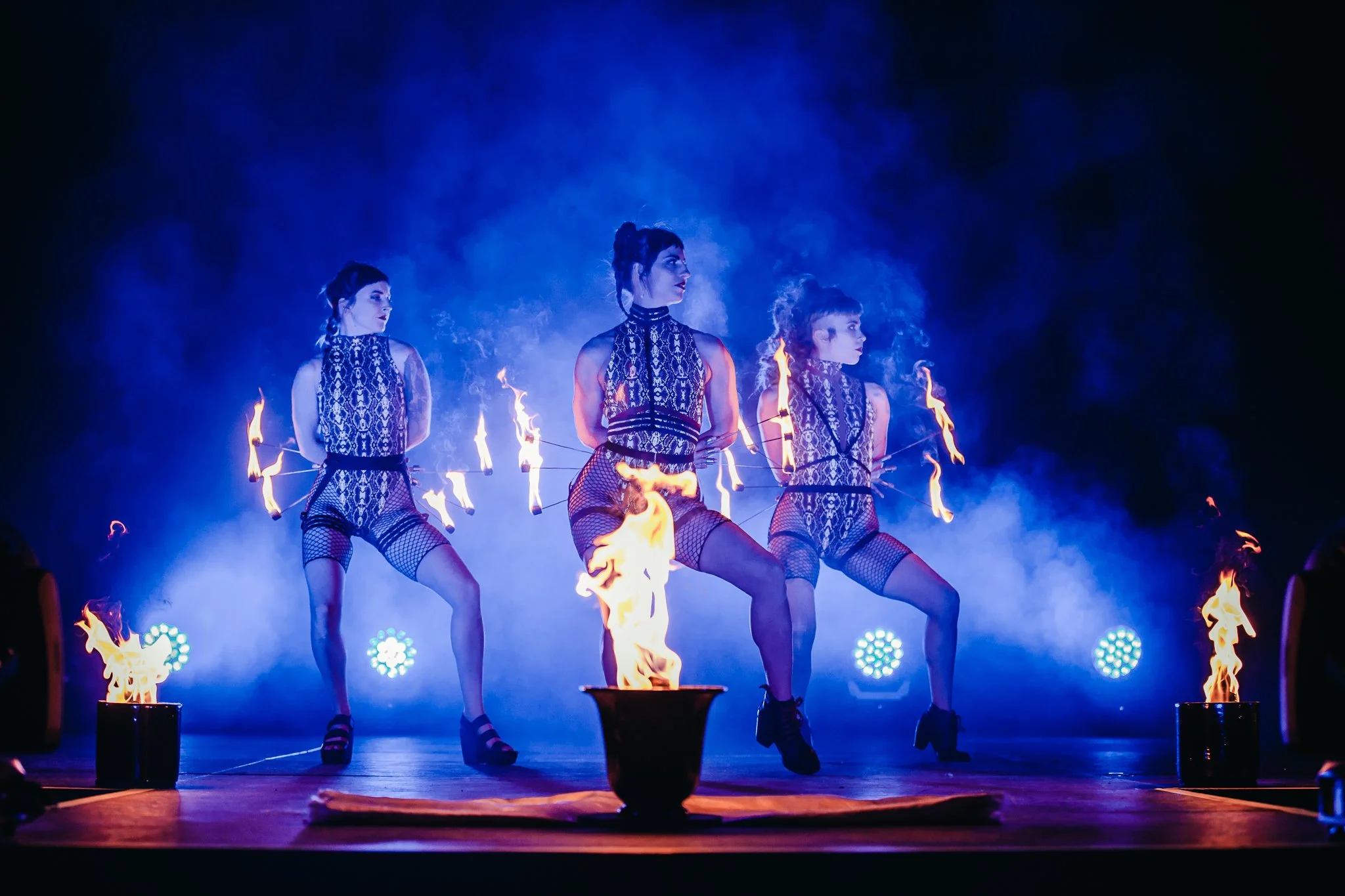 Three female performers dressed in snake print outfits perform a fire dance on a stage, with flames and blue stage lighting creating a dramatic effect.