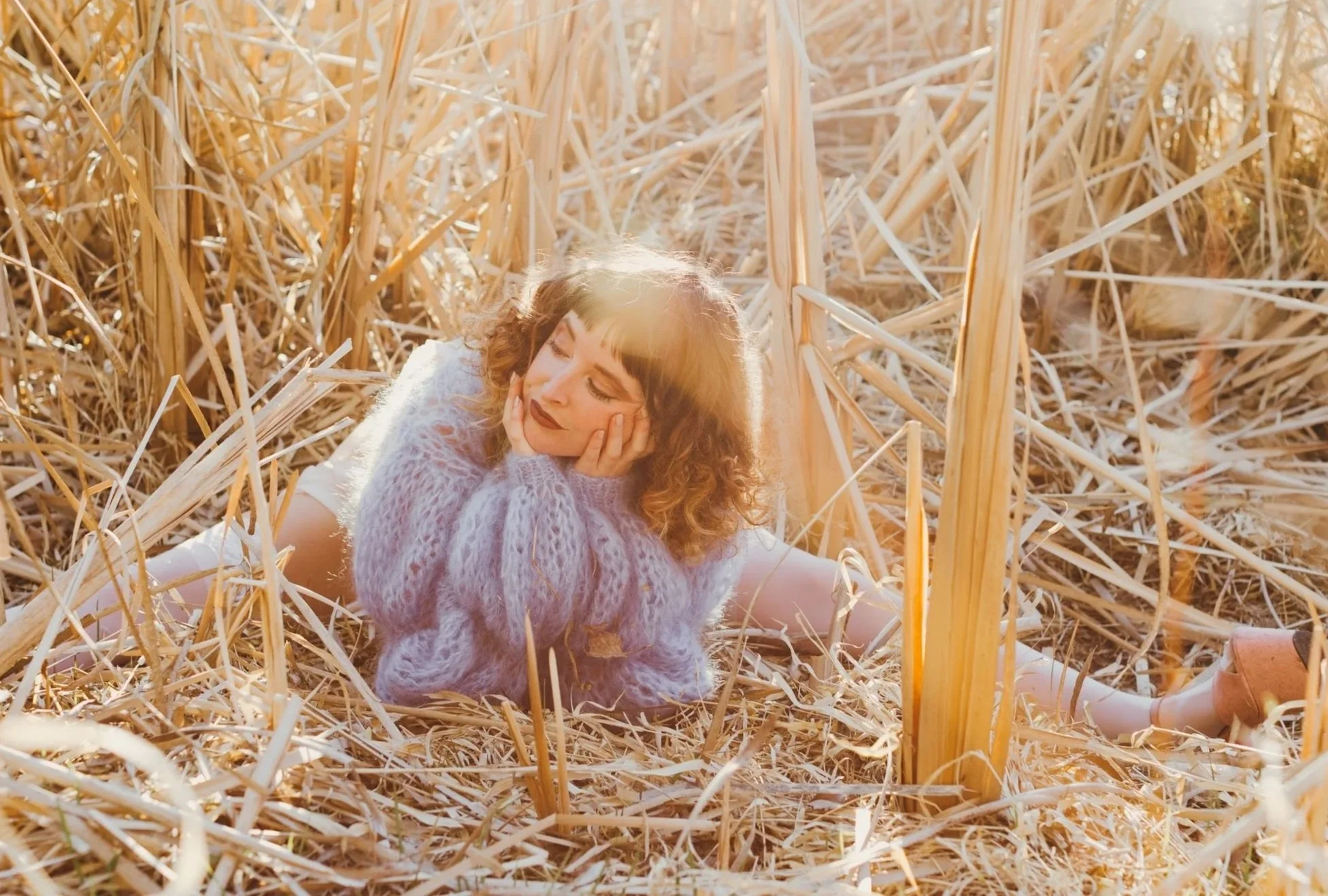 Young woman with curly hair, wearing a lavender sweater, sits among dried straw with eyes closed and hands on her face, illuminated by warm sunlight.
