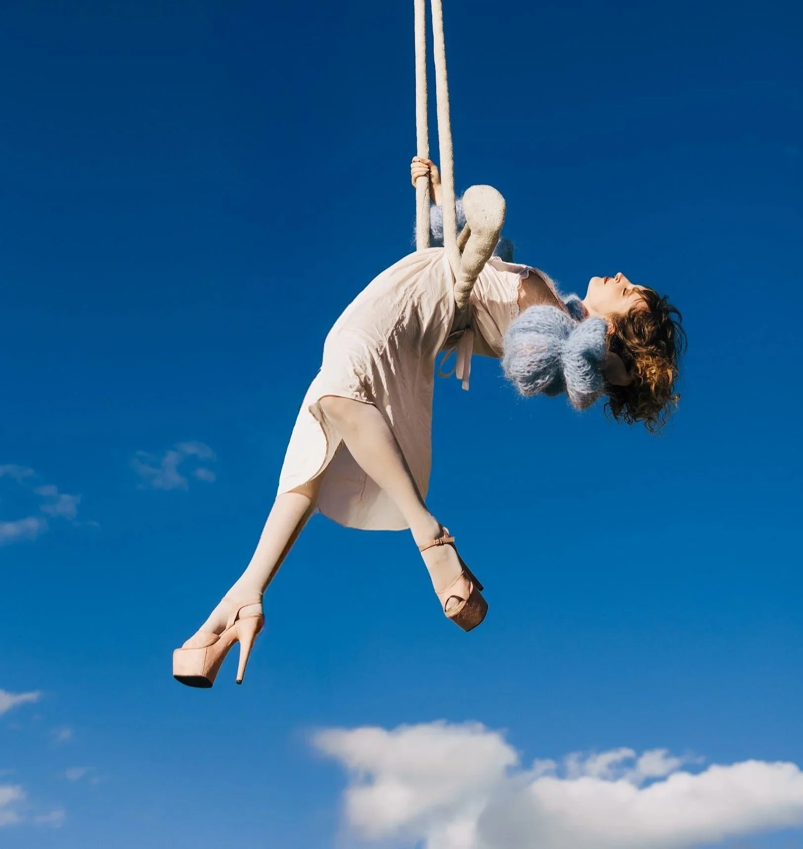A woman in high heels and a light-colored dress swinging on a rope against a bright blue sky with a few scattered clouds.