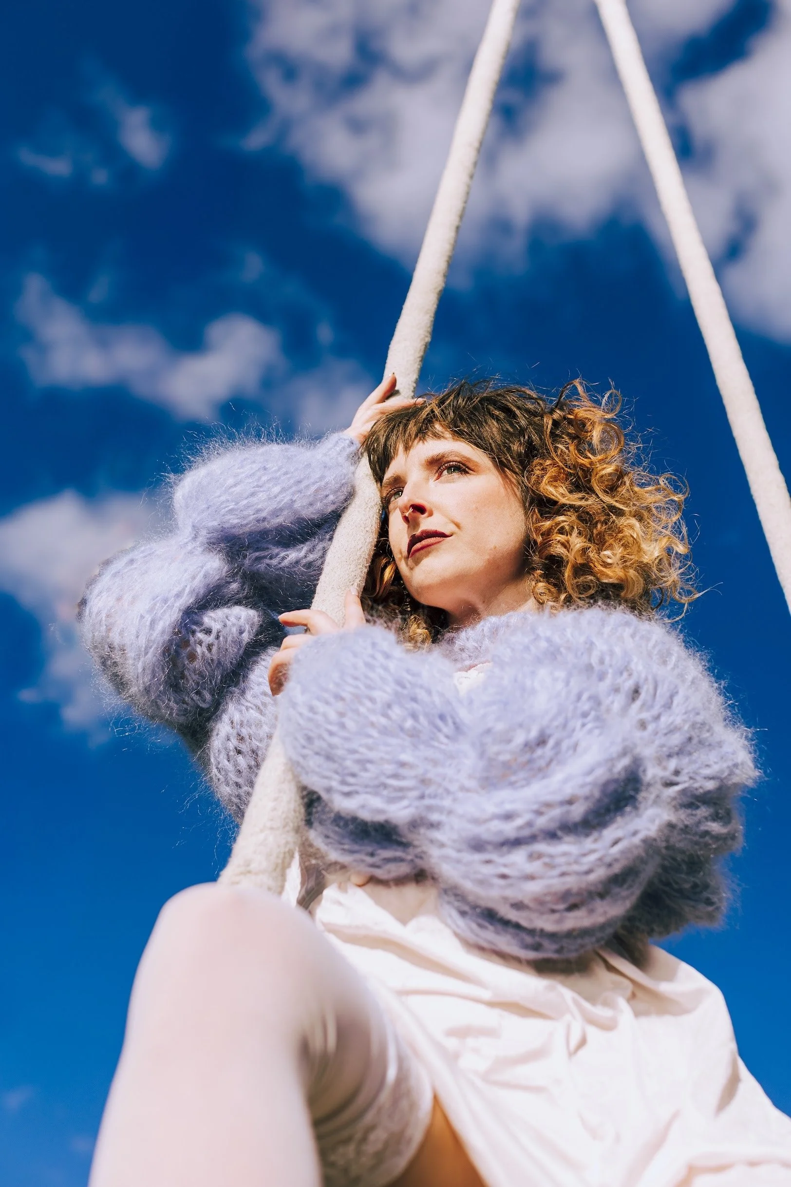 A woman with curly hair wearing a fuzzy sweater sitting on a swing outdoors against a partly cloudy blue sky.