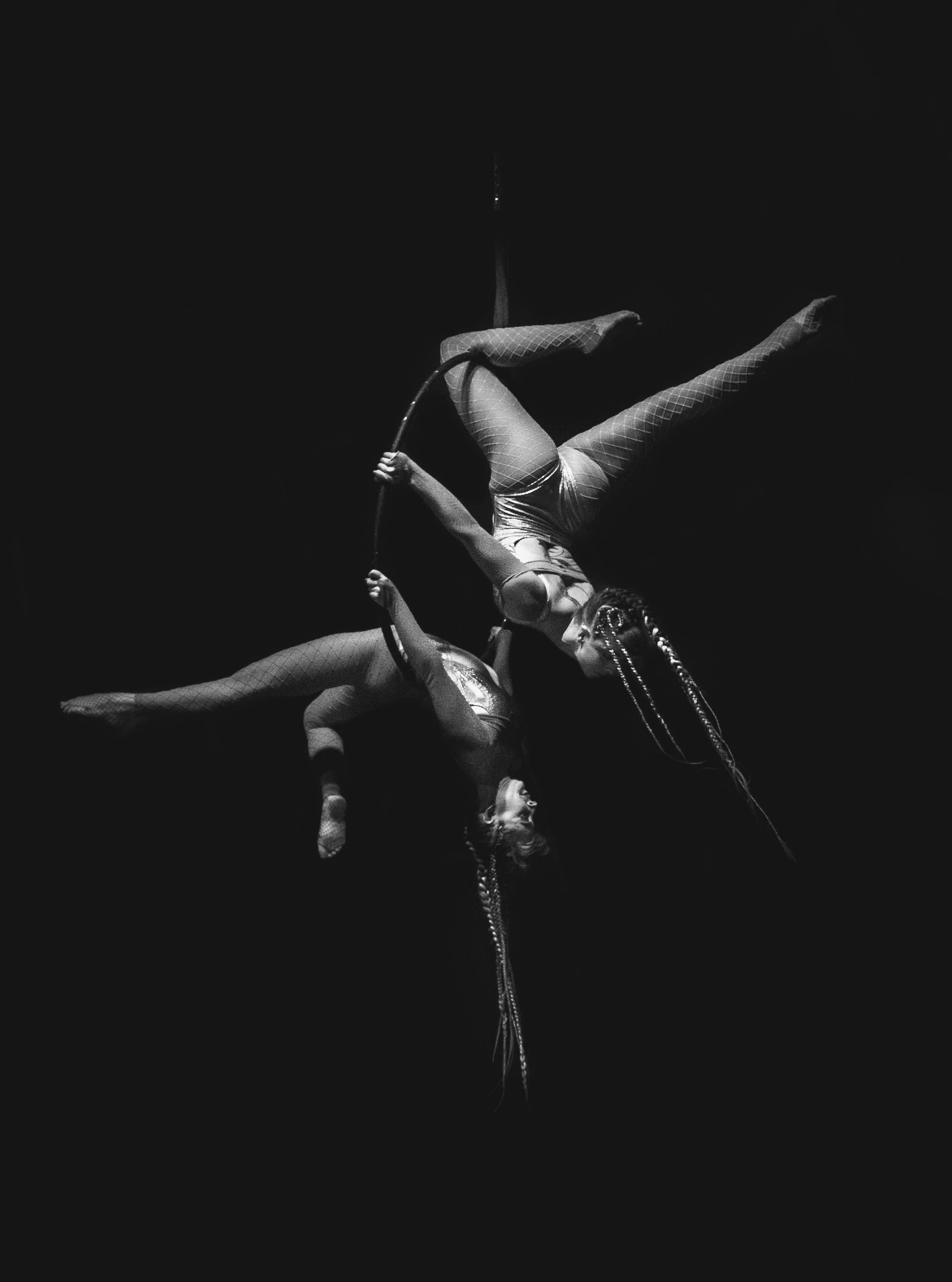 Two acrobats perform an aerial act on a hoop in a dark setting, with one hanging upside down and the other holding the hoop, both dressed in patterned outfits.