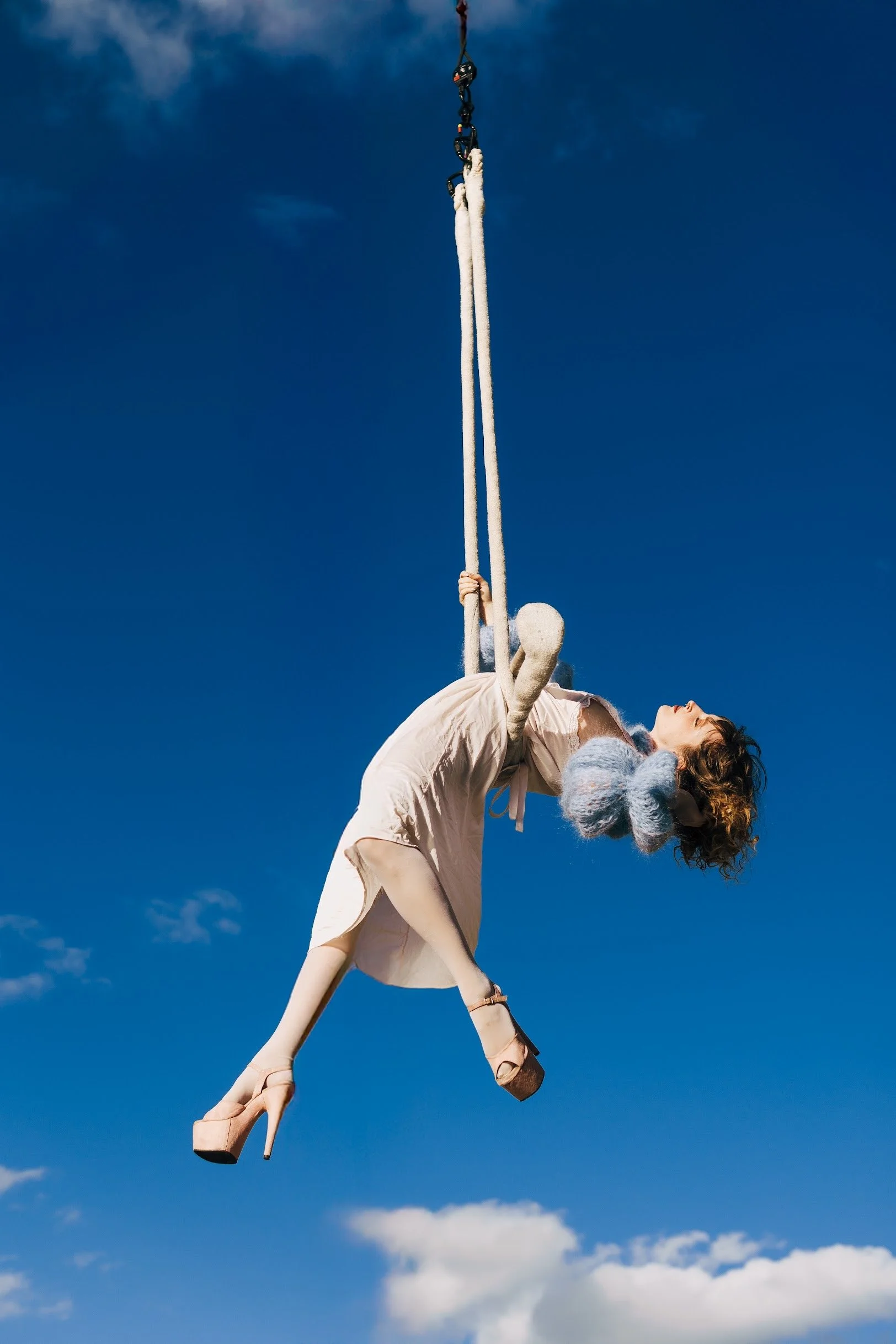 A person wearing a white dress and high-heeled shoes is hanging upside down from a trapeze against a bright blue sky with some clouds. They hold onto the trapeze ropes with their hands, and a stuffed animal is held in their arms.