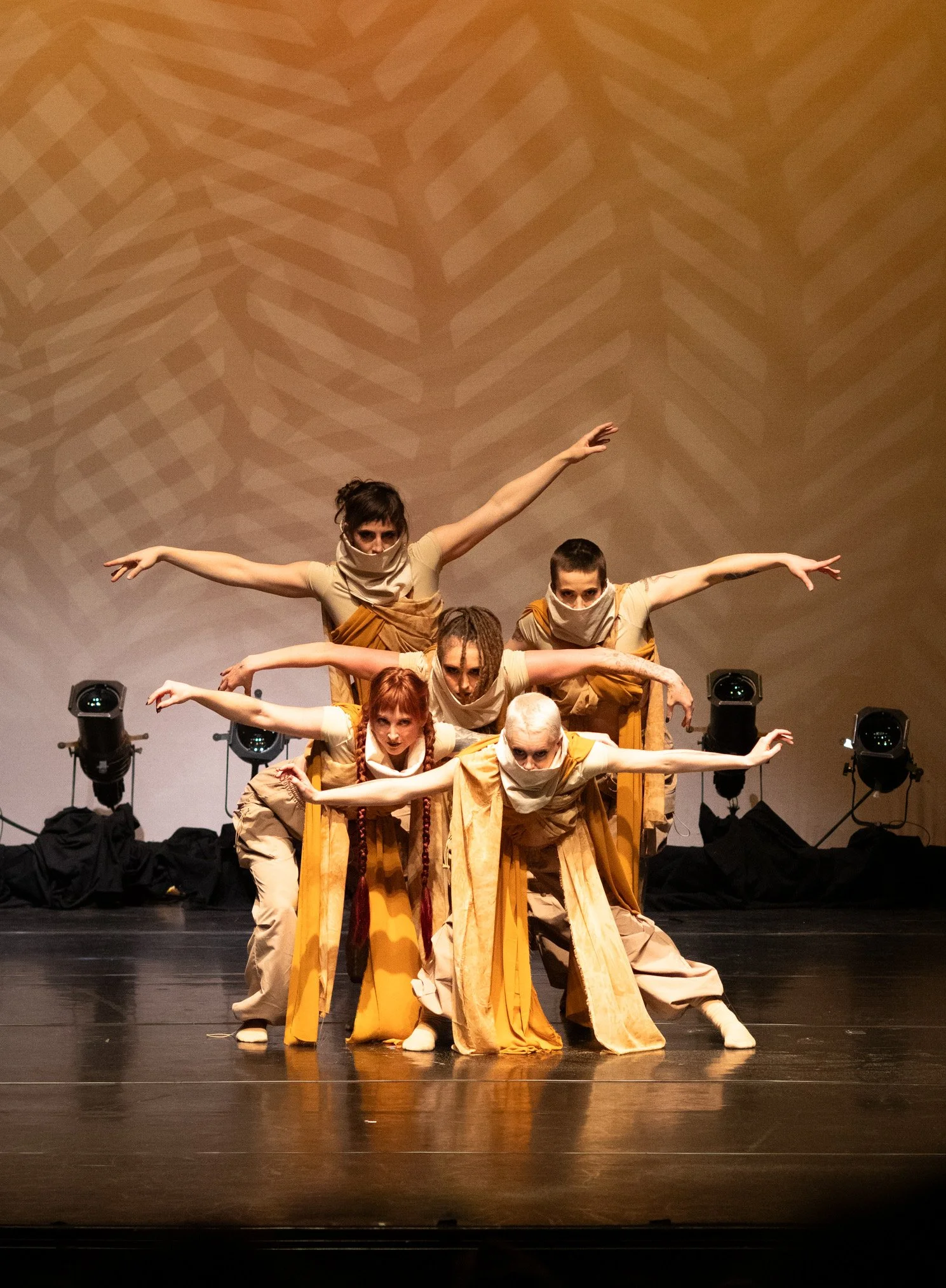 Group of seven performers in matching beige and yellow costumes on stage, striking a pose with arms extended, with a patterned background and stage lights.