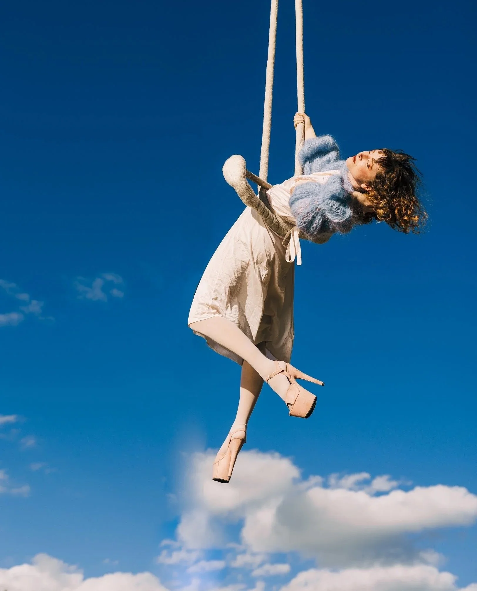 A woman with curly hair wearing a fluffy sweater and a skirt on a swing against a clear blue sky with some clouds.