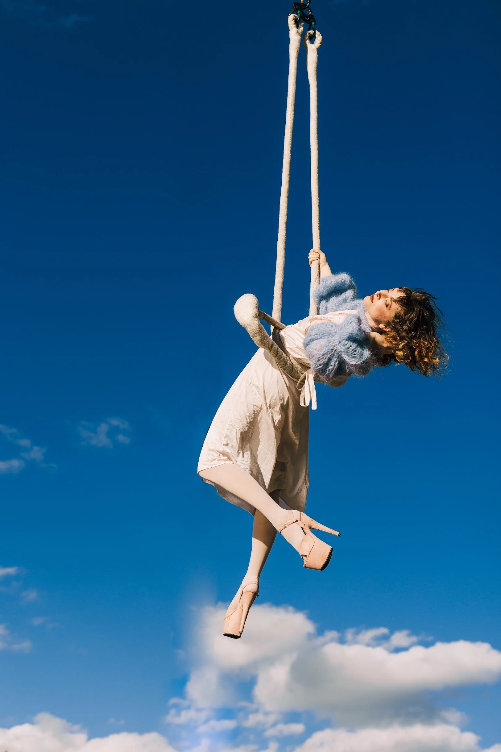 A woman dressed in vintage clothing, including high heels, tights, a skirt, a blouse, and a fluffy coat, is swinging on a large rope against a clear blue sky with a few clouds.